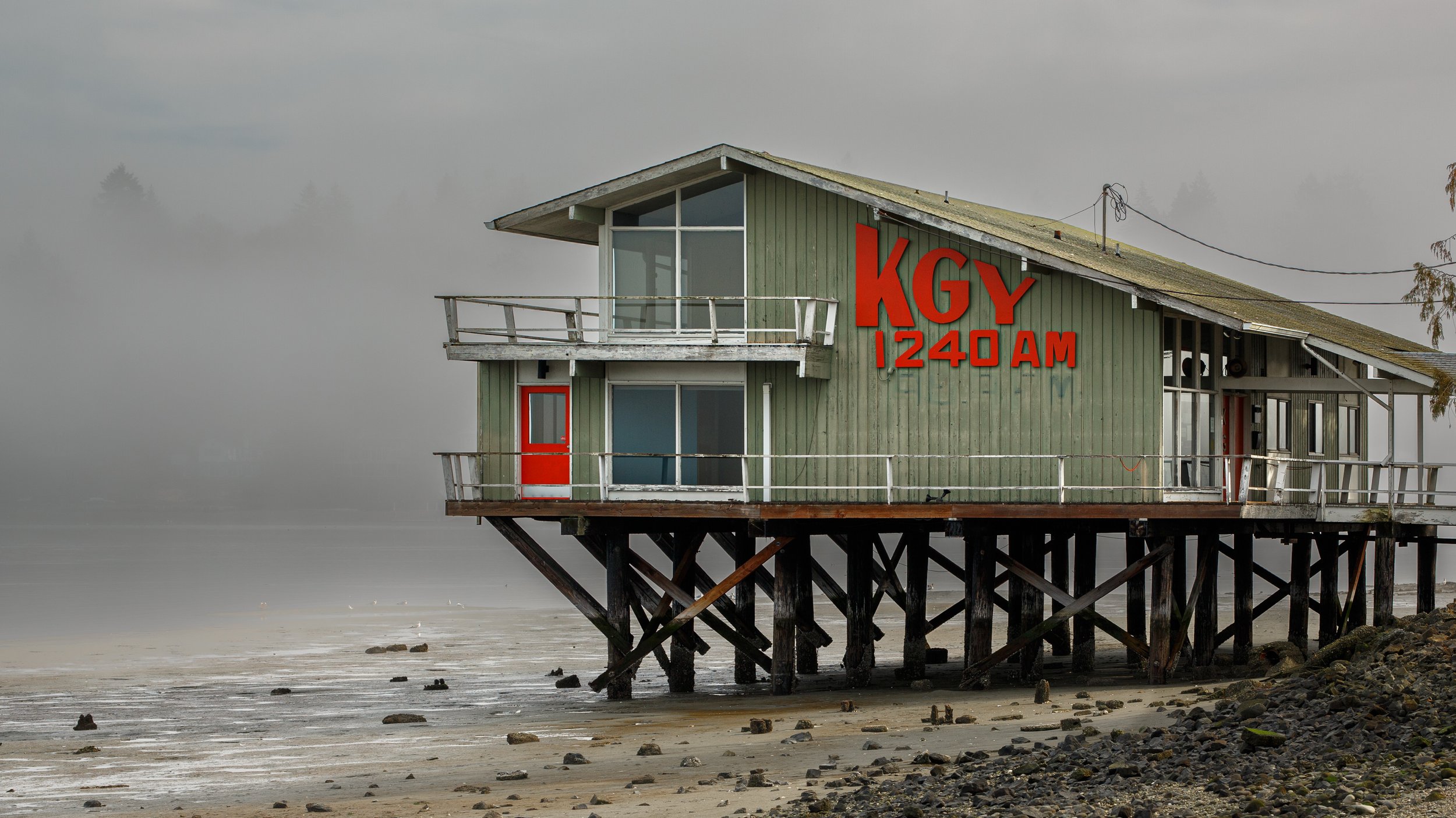 KGY 1240 AM radio station building stands on wooden pilings in Olympia Washington. Green weathered siding features red signage and door. Low tide reveals rocky shoreline under morning fog.
