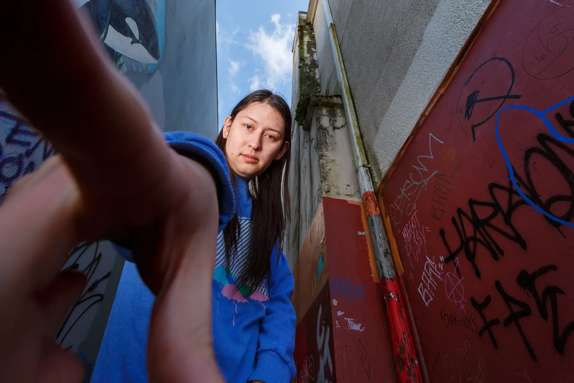 Low angle, wide-lens portrait of comedian Nat Penrose reaching her hand directly down toward the camera, standing in a downtown Olympia alleyway flanked by a red, graffitied wall.