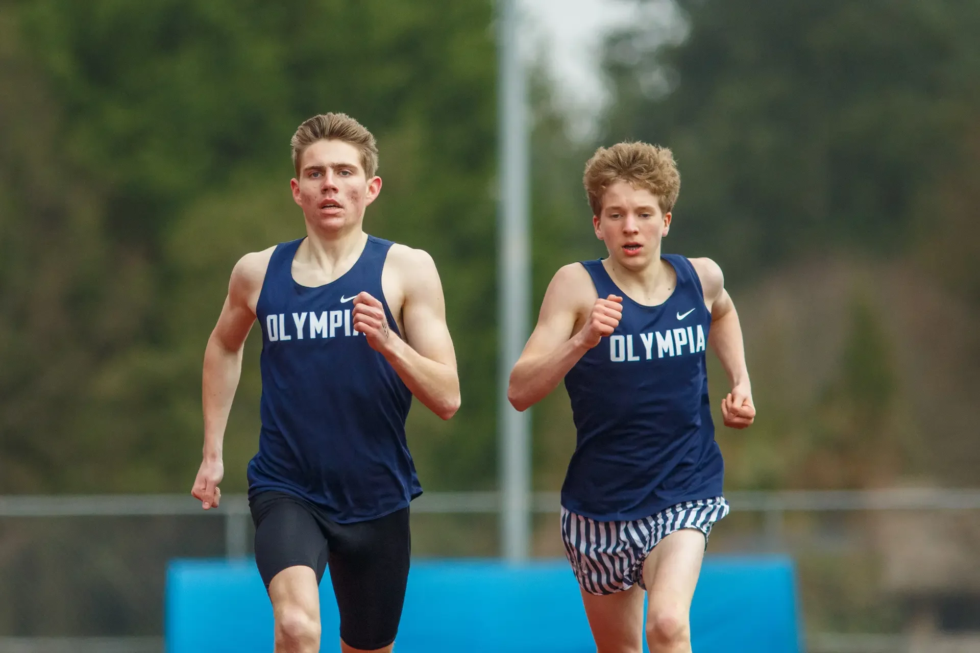 Two male high school track athletes, Caden Durocher and Quention Lanese, running side-by-side in blue uniforms during a race at Olympia High School, photographed by Russell Moore.