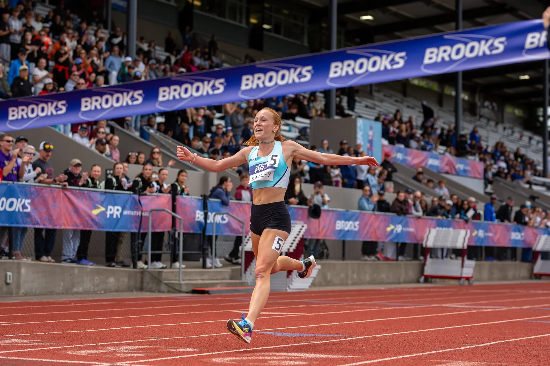 Chiara Dailey celebrating with her arms raised just before breaking the finish line tape to win the Girls 2 Mile at the 2026 Brooks PR Invitational in Renton, WA.