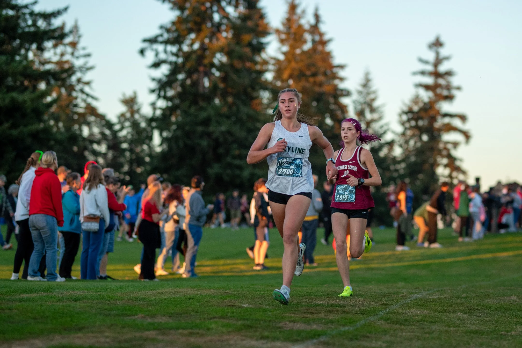 Skyline runner Lucy Fraser in a white jersey and Northwest School runner Maggie Hurtado in maroon running together across the golf course.