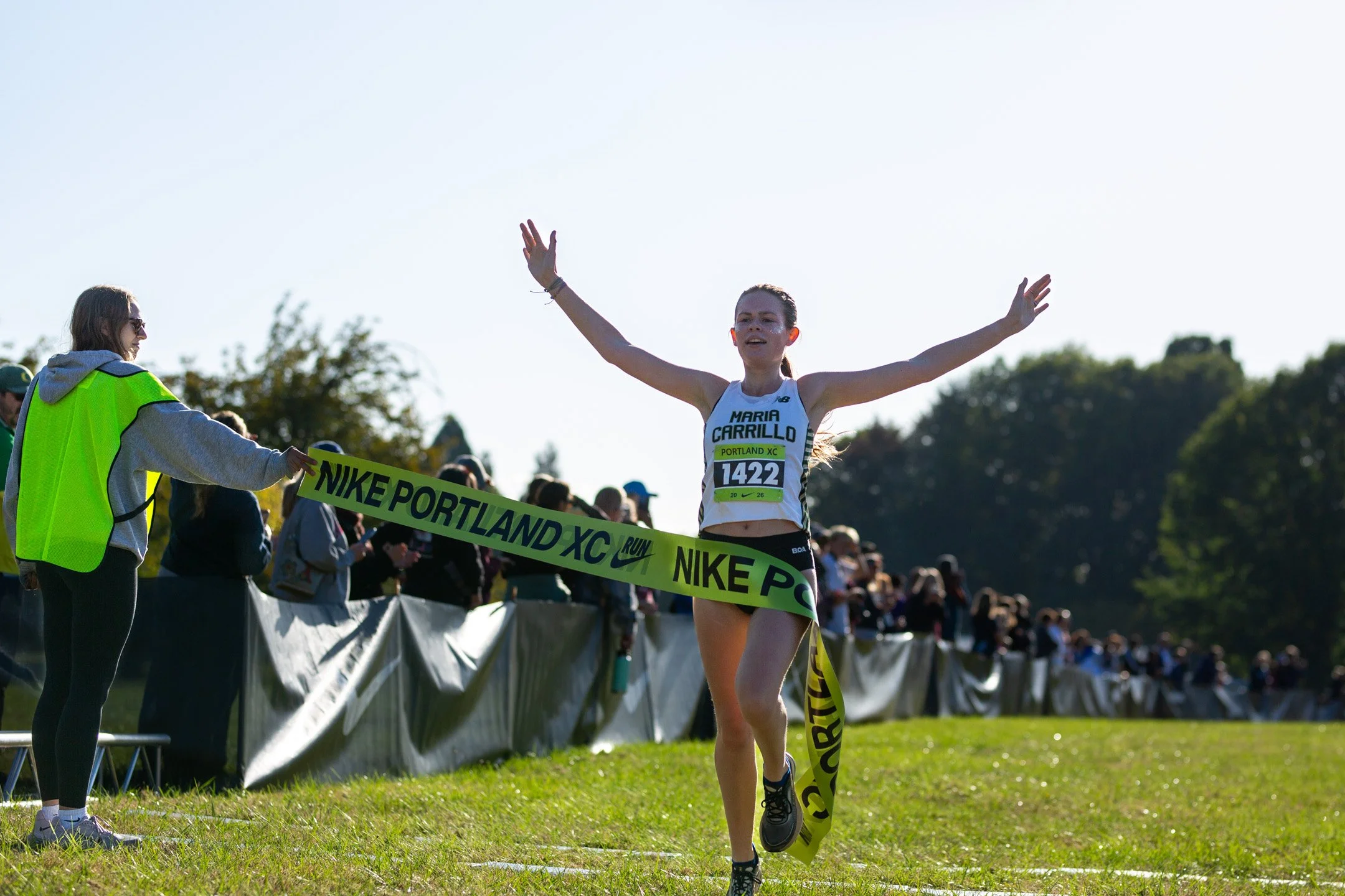 Maria Carrillo's Joslyn Tribbey raises both arms as she breaks the Nike Portland XC finish tape held by a volunteer in a green vest.