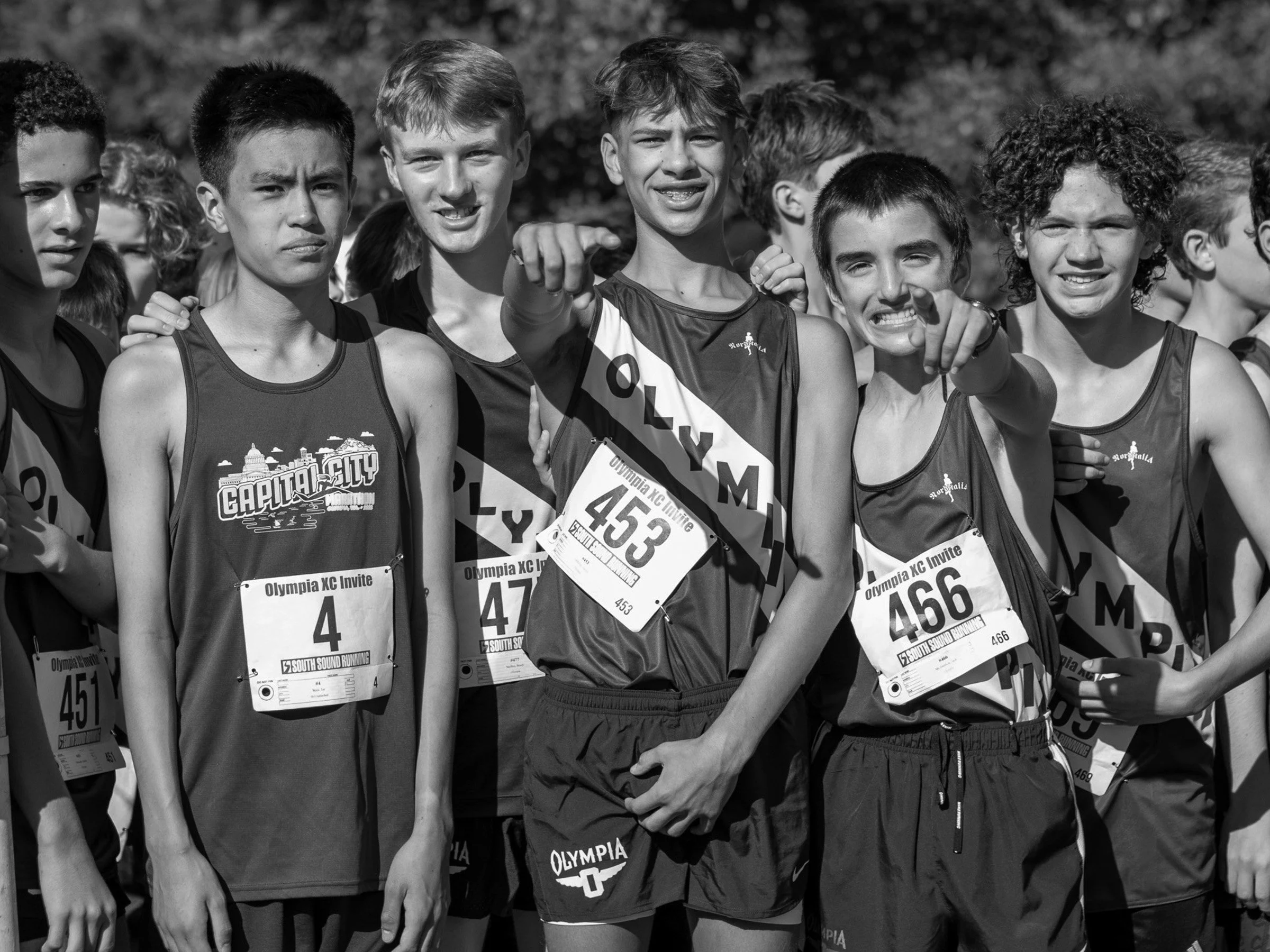 Young cross country runners in Olympia singlets smiling and pointing at the camera before a race.