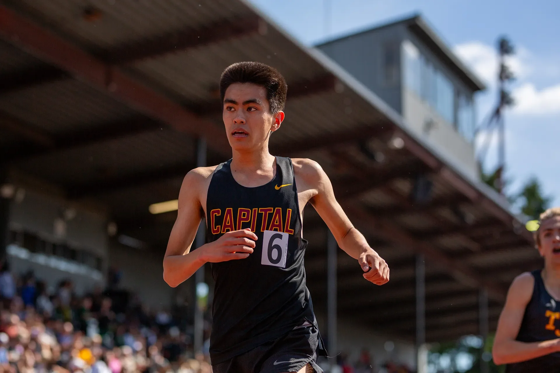 Capital High School runner Jae Walz competing in the boys 1600m race, wearing a black singlet with red lettering, with the French Field grandstands blurred in the background.
