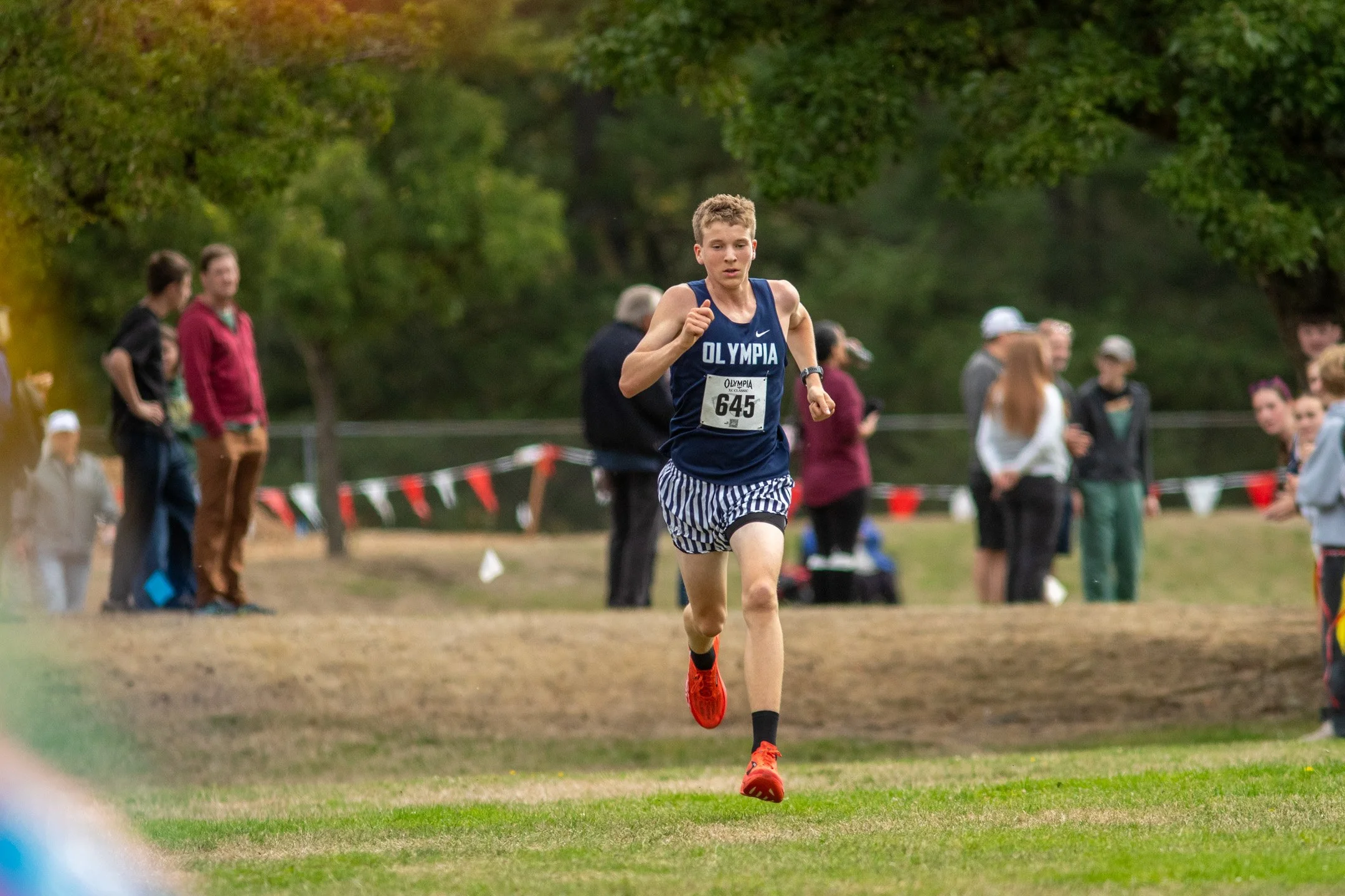 Boy in navy Olympia jersey, bib 645, running alone along a cone-lined course with autumn trees in the background.