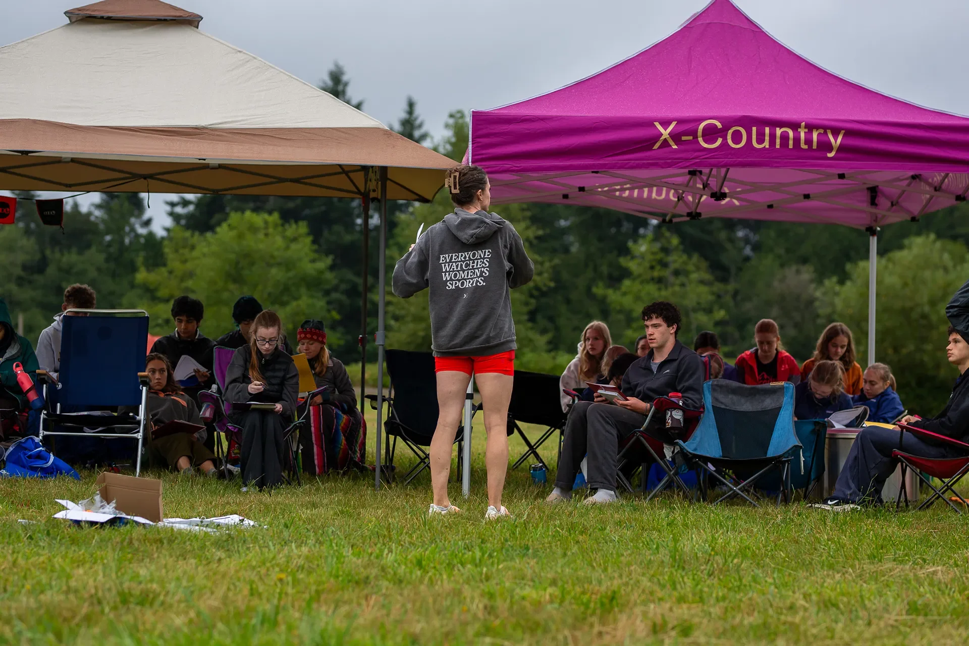 Jena Winger wearing a Togethxr hoodie stands and speaks to a group of athletes sitting in camp chairs under a pink North Kitsap cross country tent.