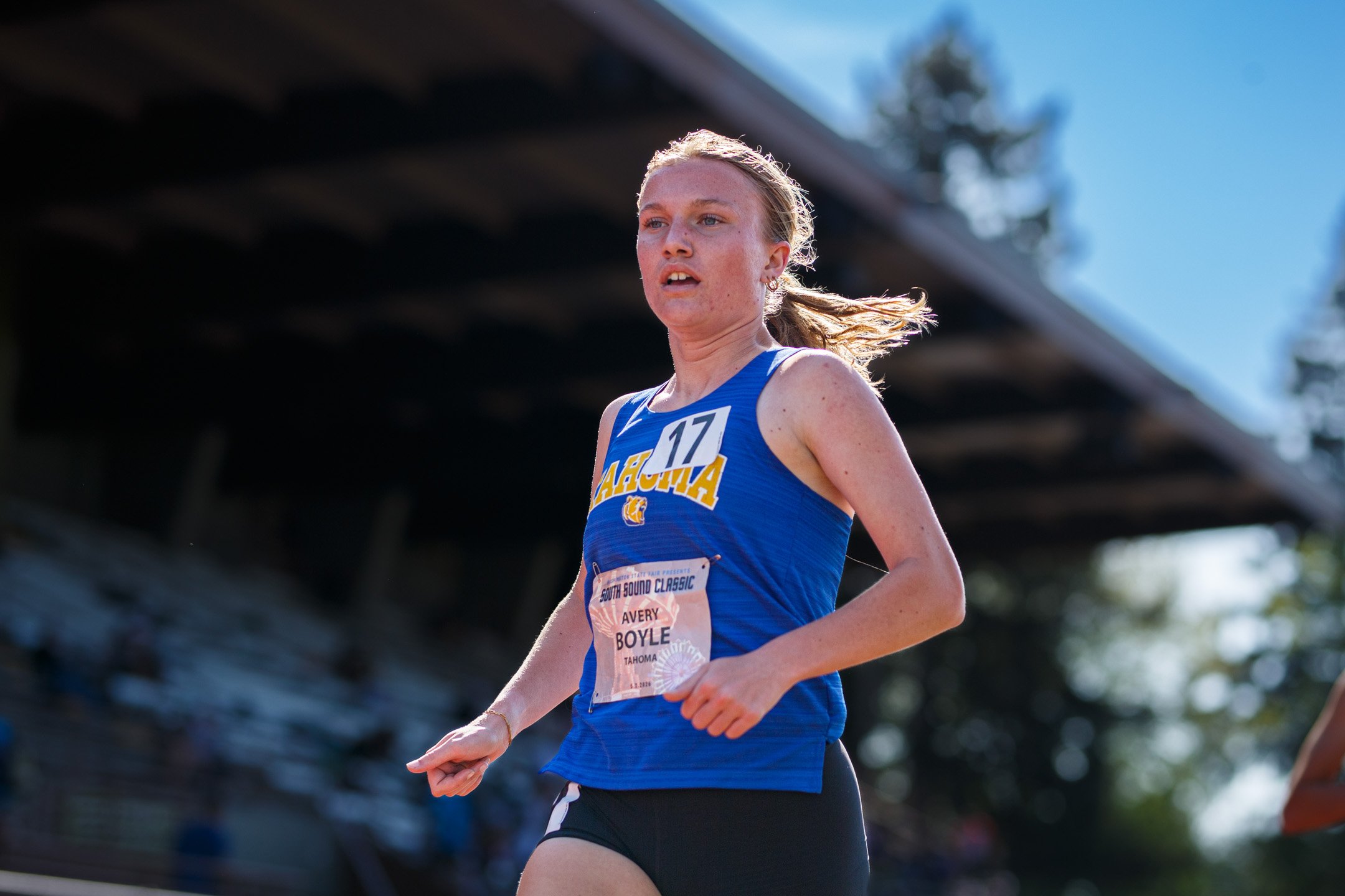 Tahoma senior Avery Boyle, bib 17, in a blue Tahoma singlet and black shorts, running in front of a stadium grandstand.