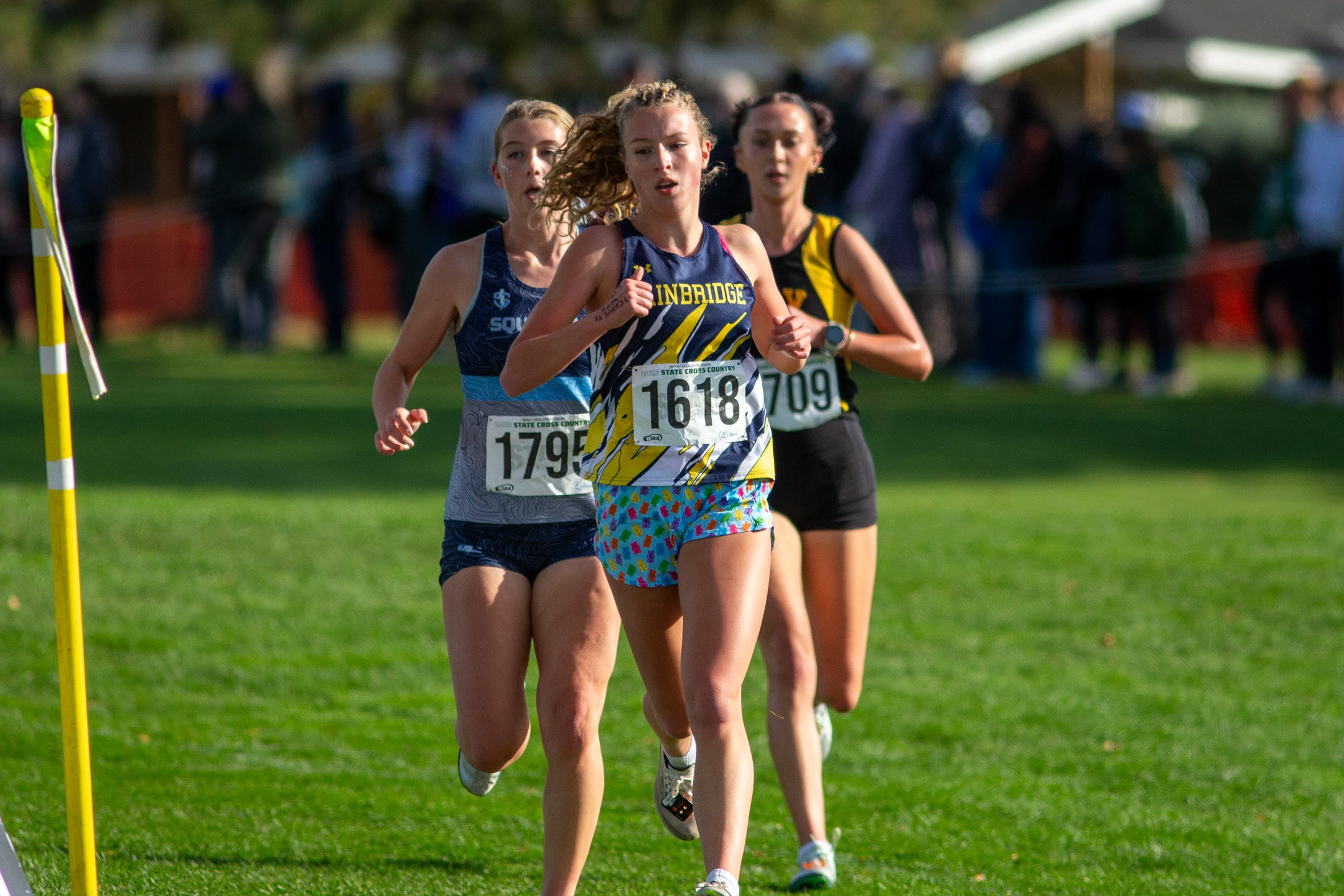 Evelyn Shutske of Bainbridge, bib 1618, runs on grass between two other runners with spectators blurred behind.