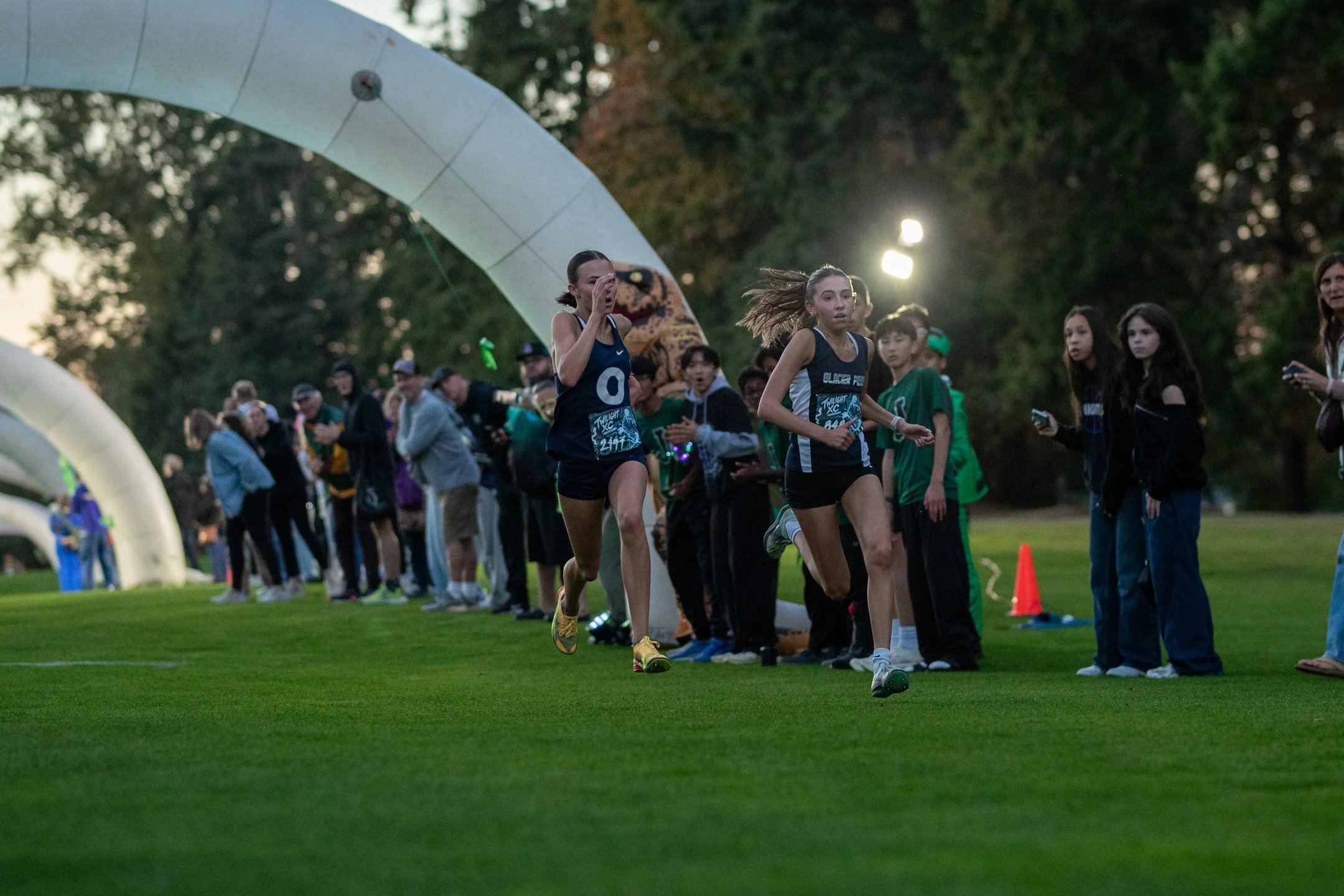 Two high school runners, Jaya Holt and Leah Hennen, sprinting neck-and-neck toward lighted white arches in the evening twilight with spectators cheering.