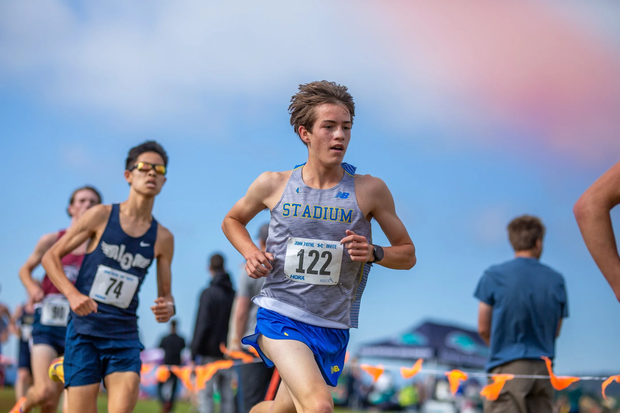 Stadium junior Will Cramer, bib 122, in a gray Stadium singlet and blue shorts, running with a runner in a navy singlet, bib 74, close behind him.