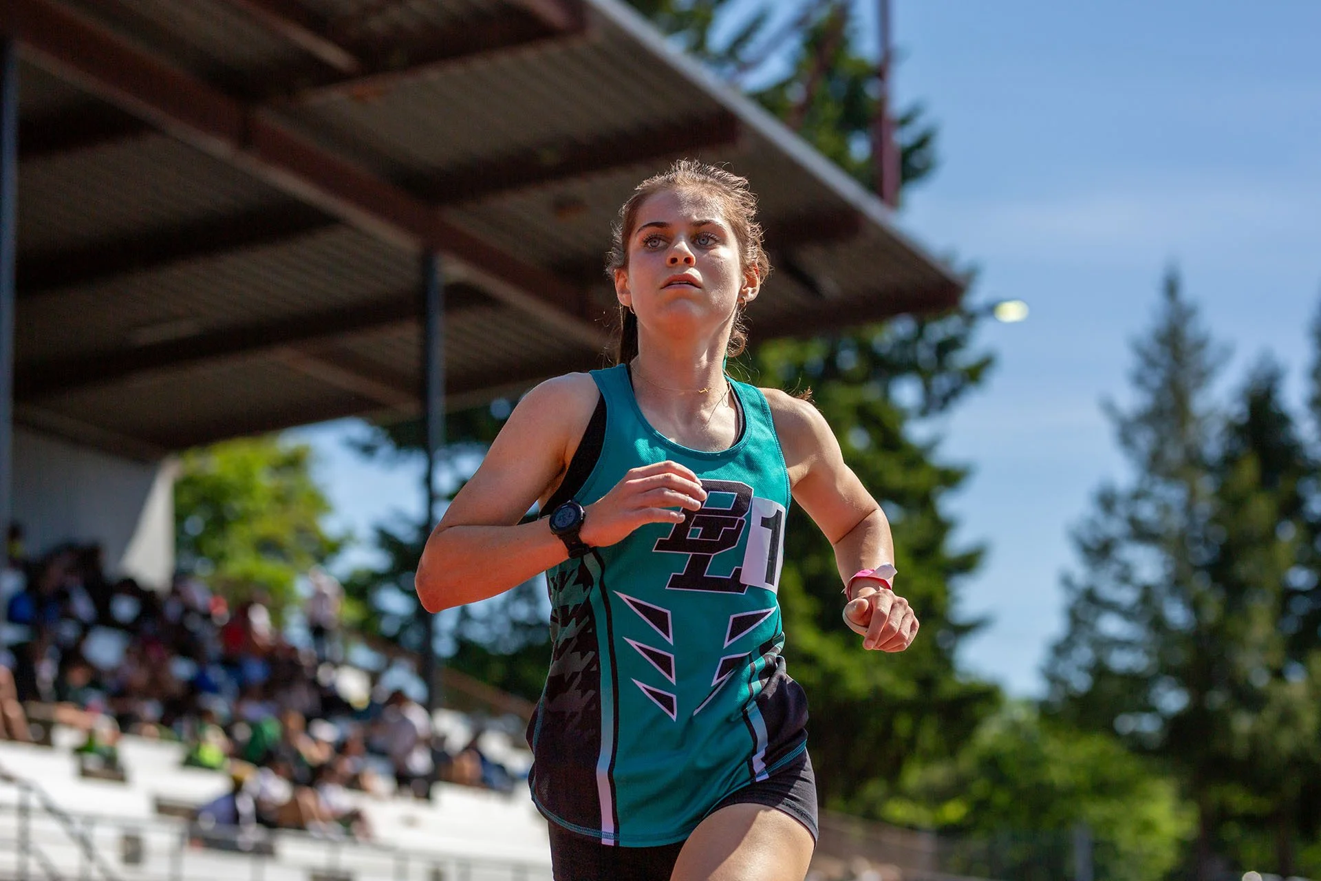Bonney Lake high school runner Latham West in a teal and black singlet, running solo on the track with blurred grandstands in the background during the 4A 3200m race.