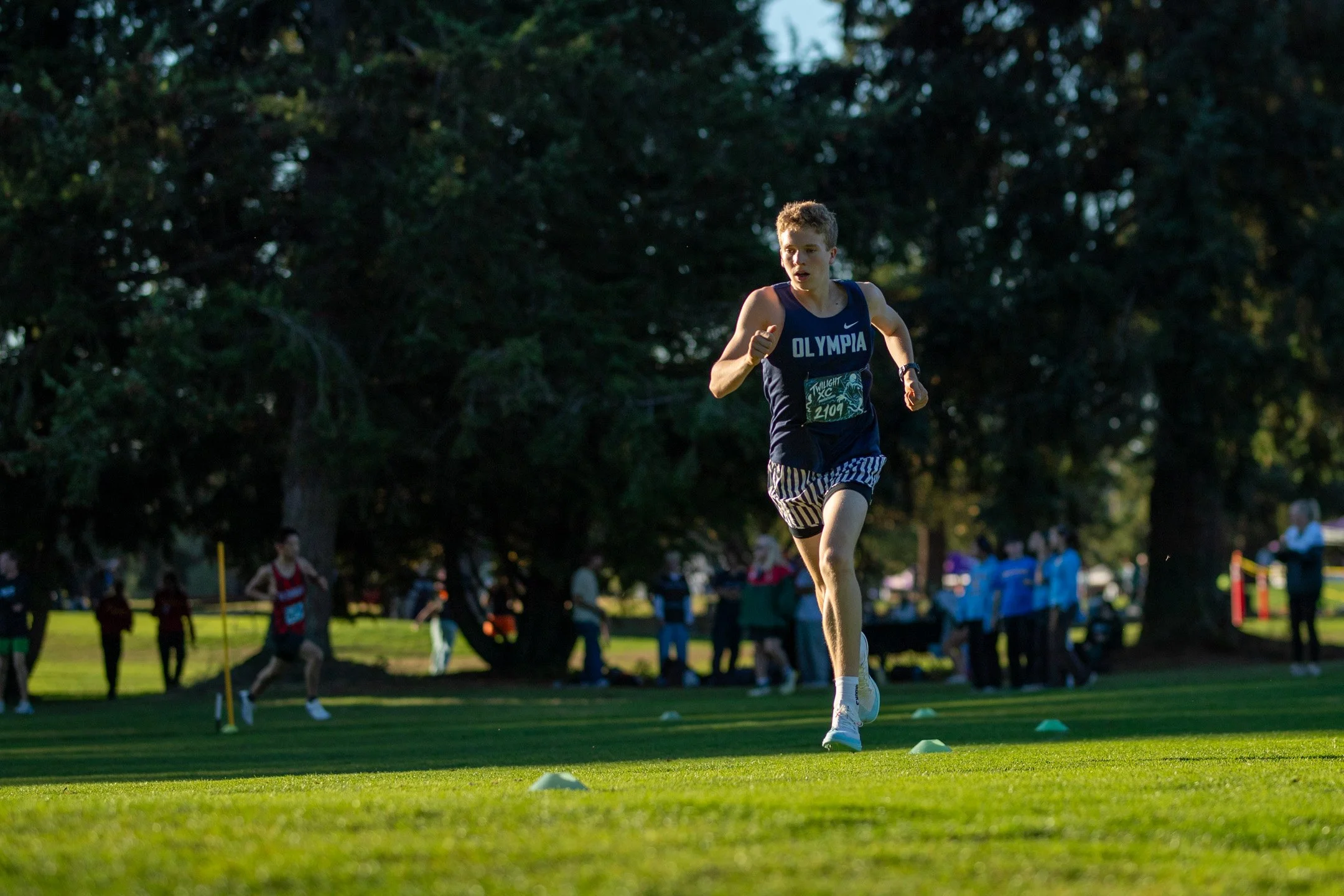 Olympia runner Quenton Lanese in a navy jersey running alone across a sunlit grass fairway toward the camera.