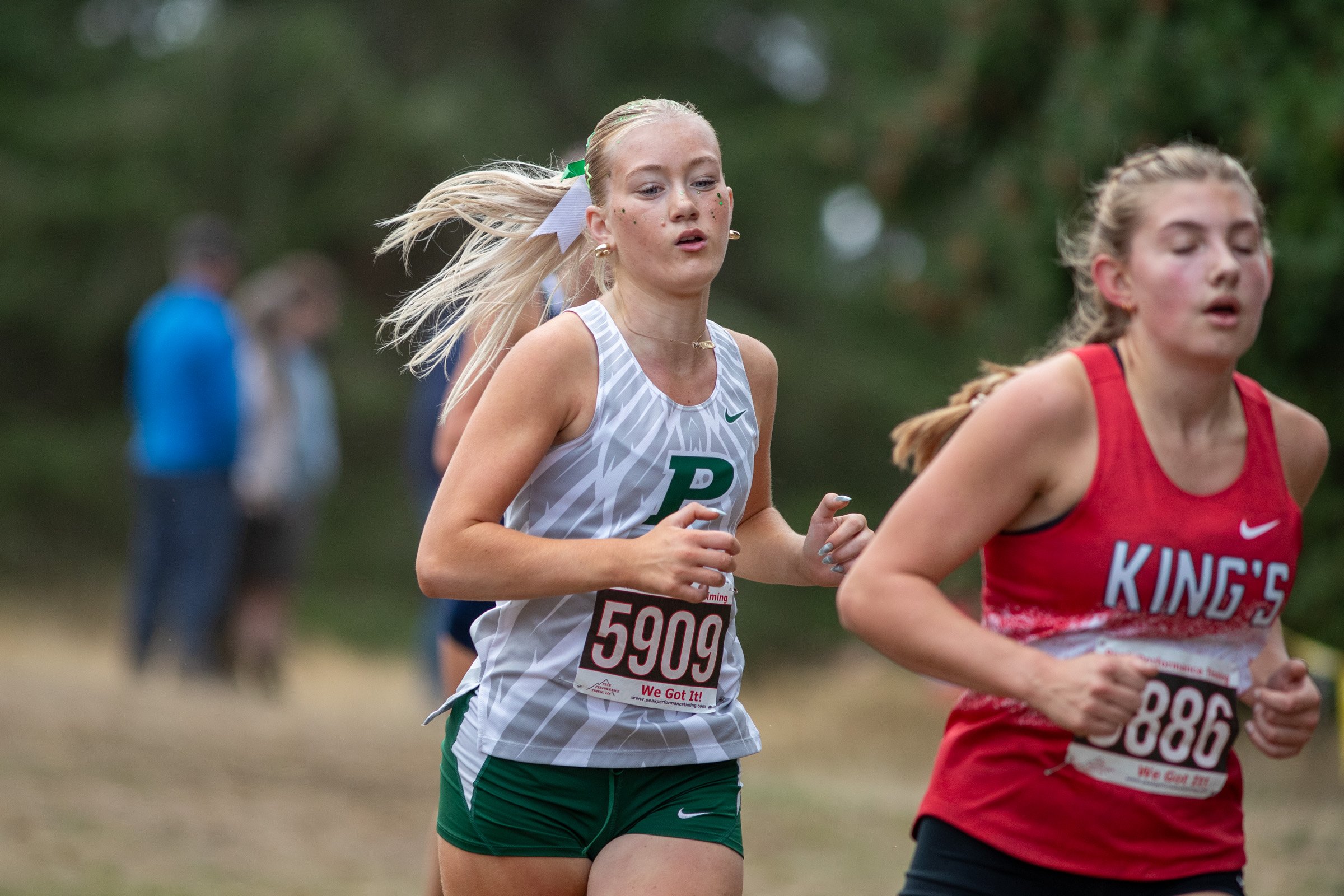 Peninsula junior Ascher Lascheck, bib 5909, in a white Peninsula singlet and green shorts, running beside a runner in a red singlet.