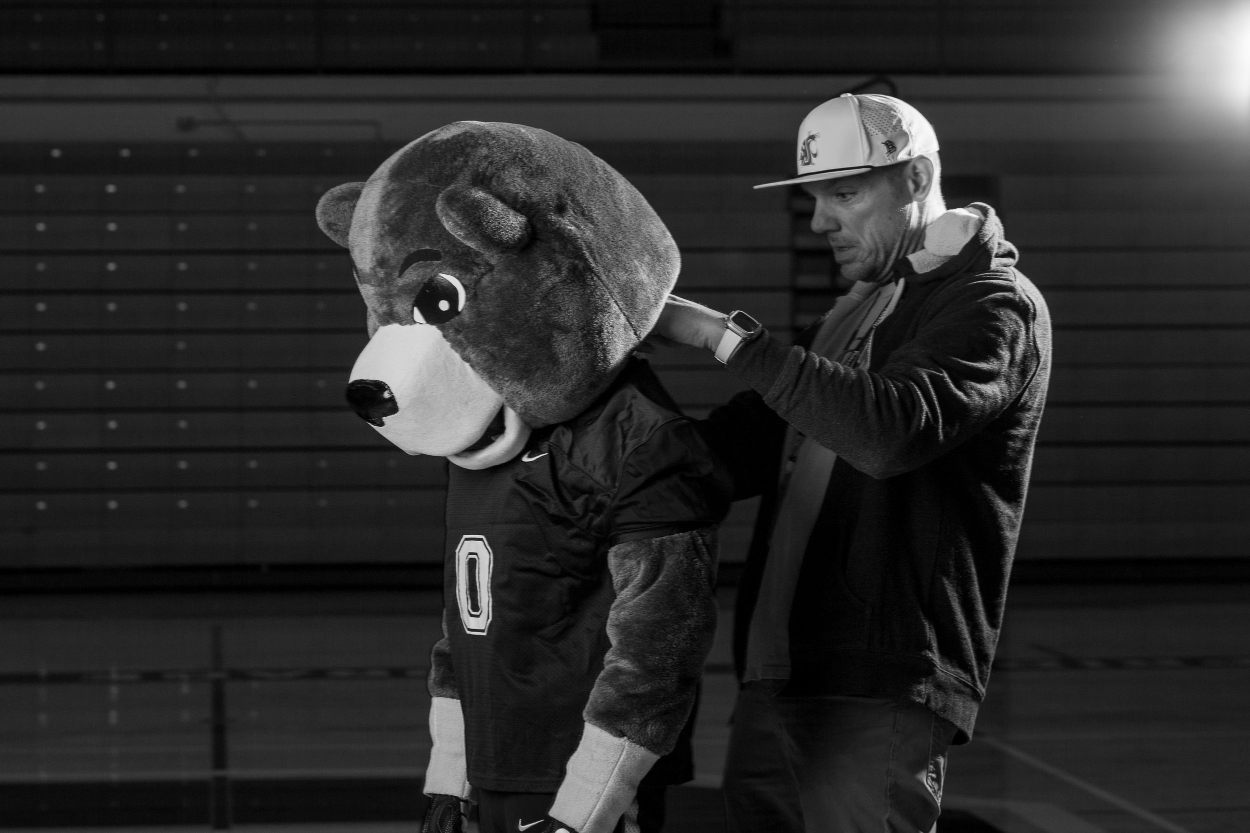 Behind the scenes black and white photo of Ryan Gerrits adjusting the head of Pepper, the Olympia High School Bear mascot, during a studio photoshoot.