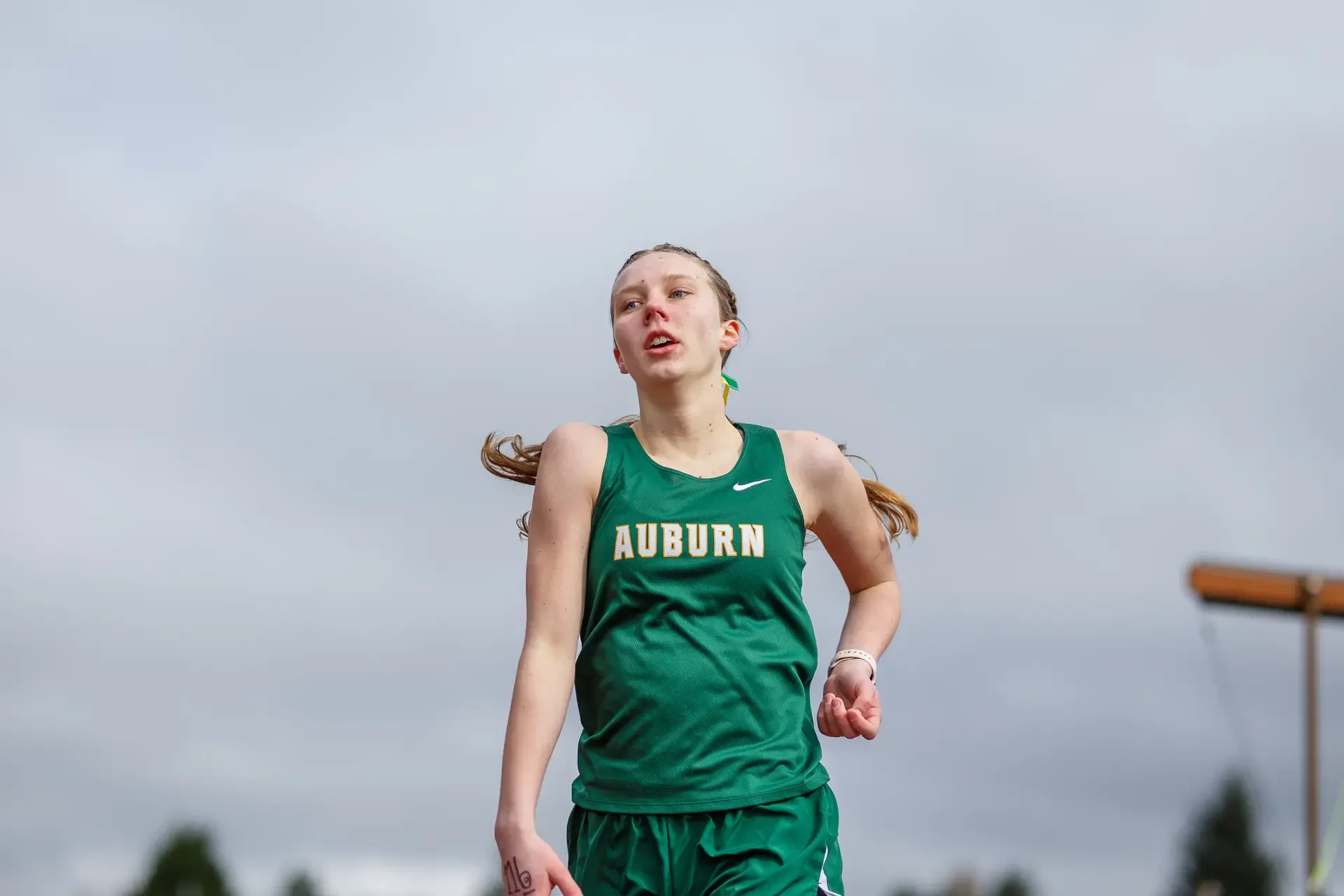 A female high school track athlete in a green Auburn jersey runs during the 1600m race at Olympia High School in Olympia, WA, photographed by Russell Moore.