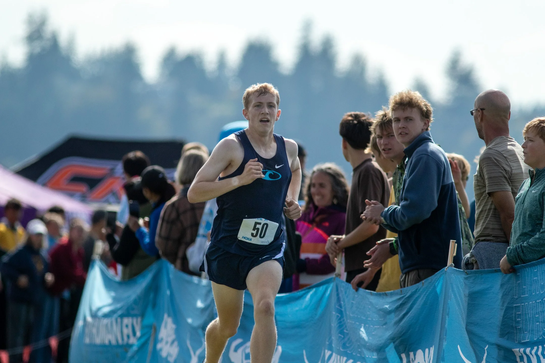 Gig Harbor runner Joshua Minner in a navy singlet with bib 50 pushing the pace down the final straightaway lined with blue banners.
