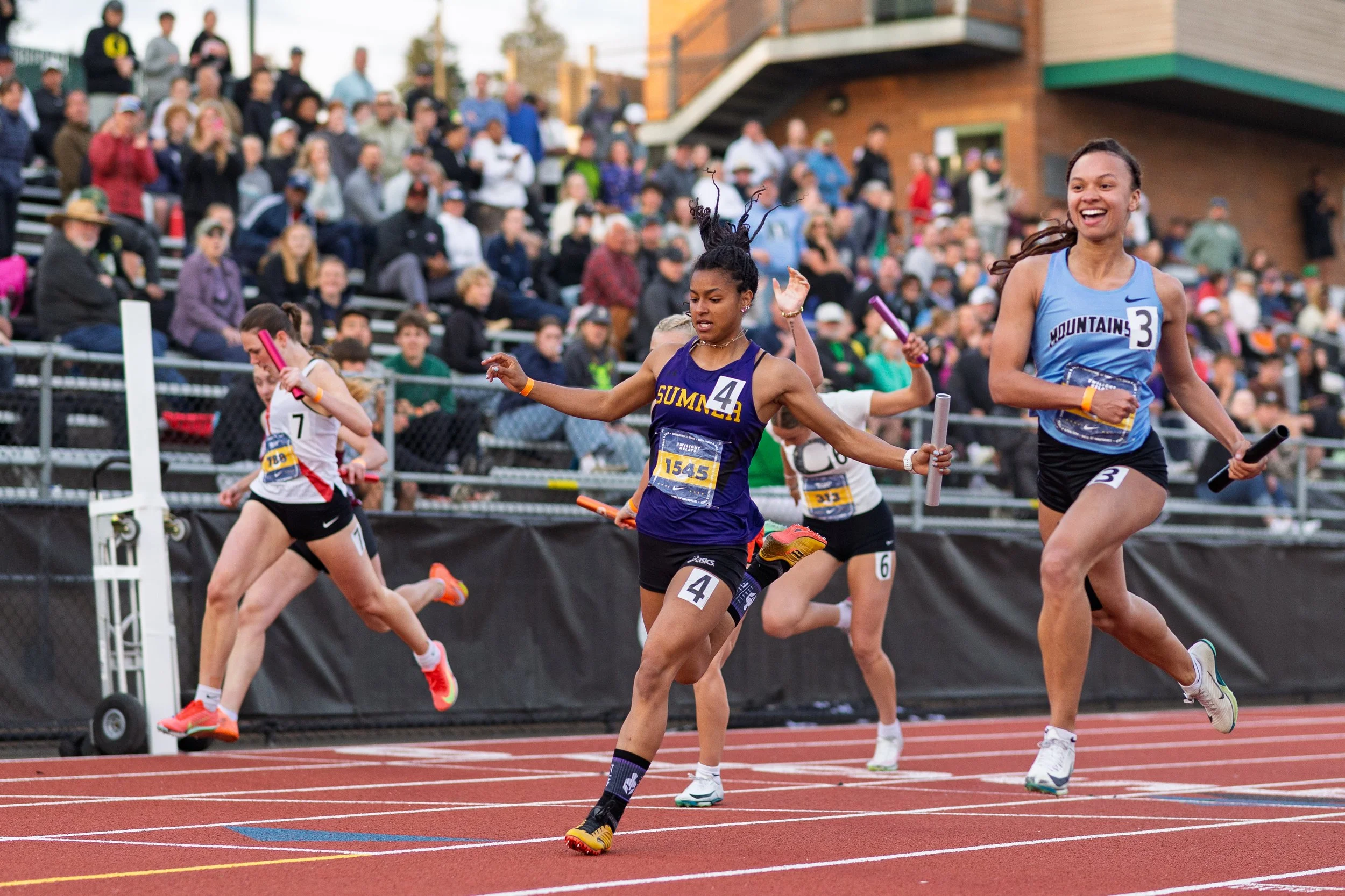 A group of female high school track athletes sprinting across the finish line with batons during the Nike Jesuit Twilight Relays, photographed by Russell Moore.
