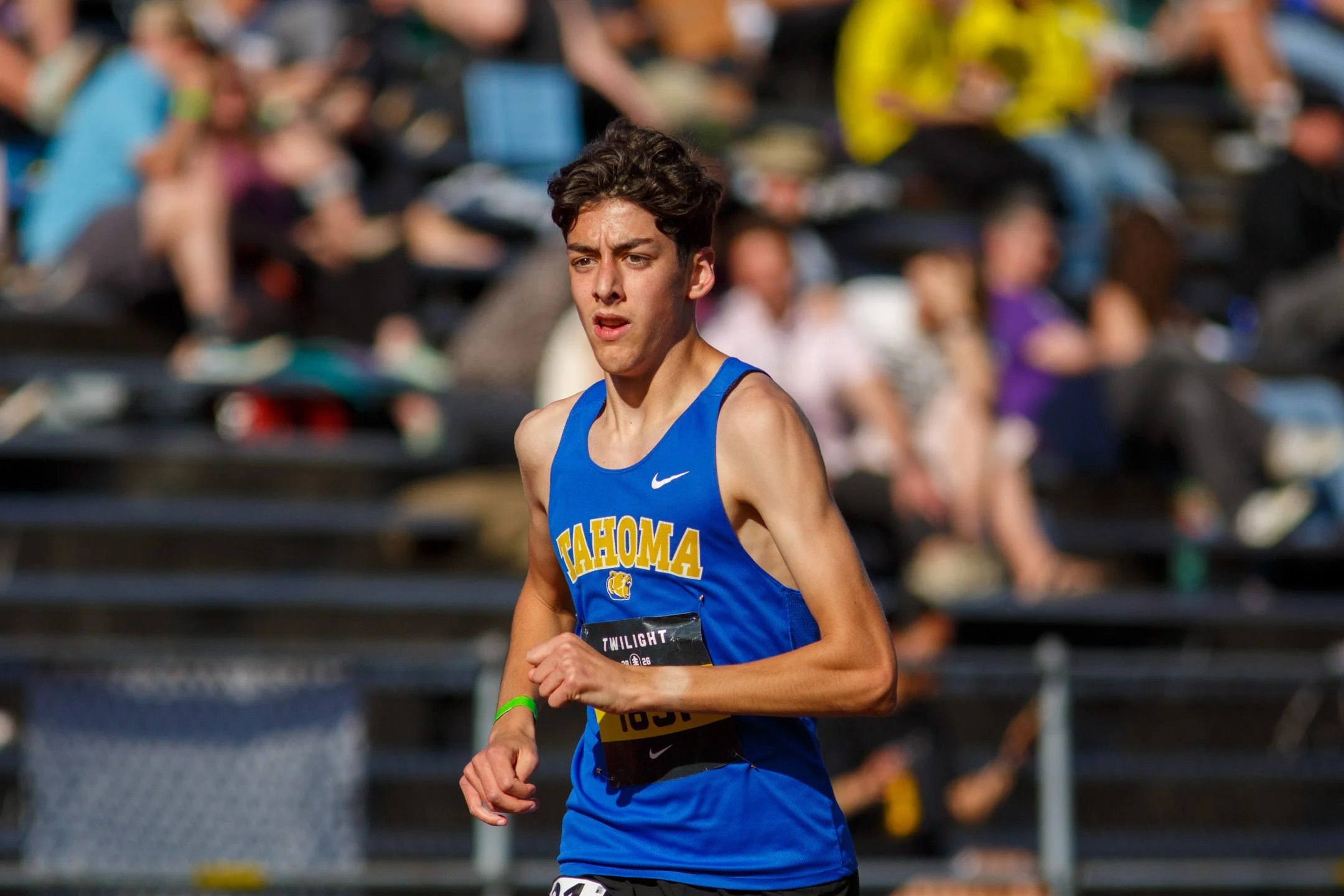 Tahoma junior Thane Winters in a blue Tahoma singlet with a green wristband, running past spectators in the stands.