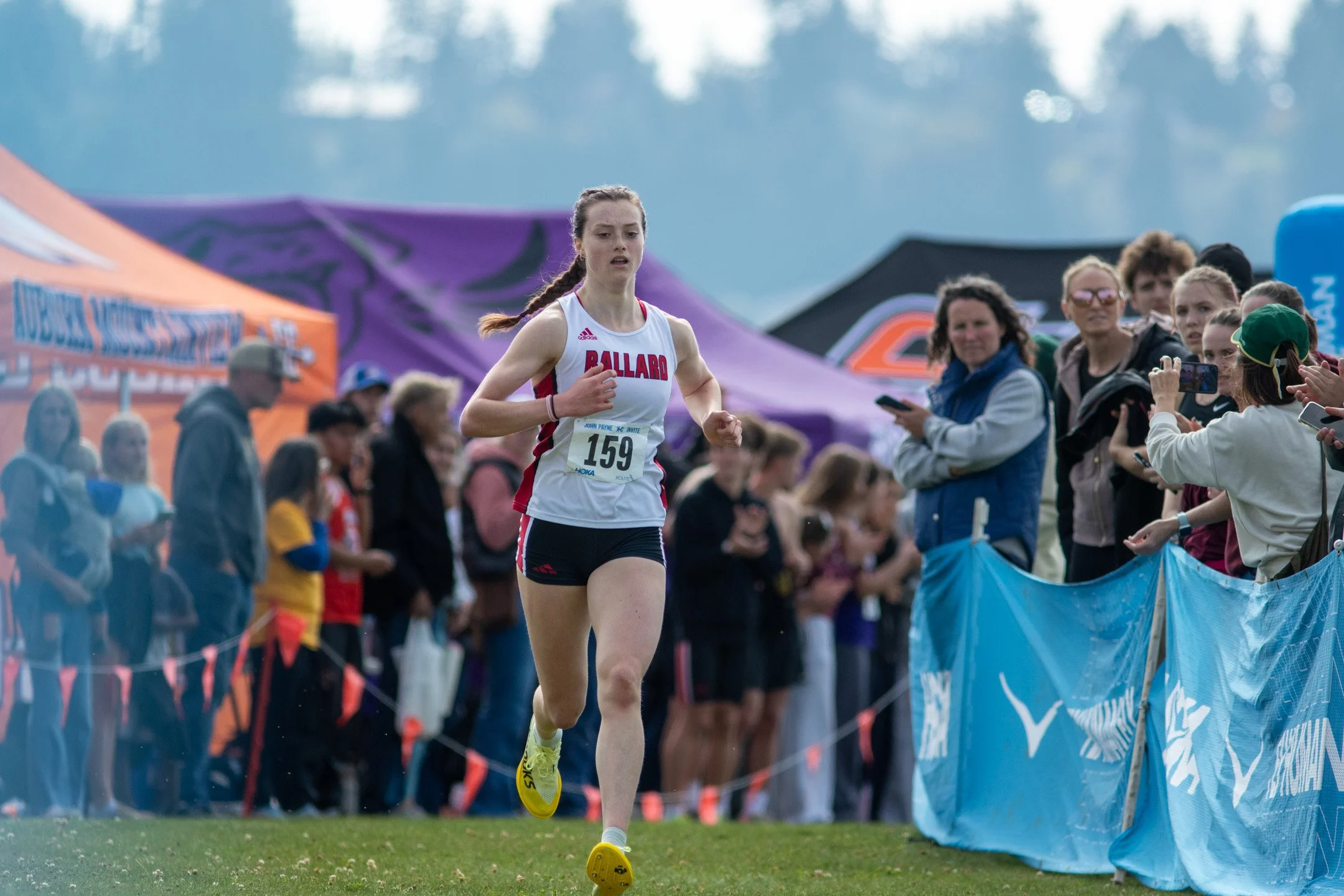Ballard High School runner Ciana Johnson in a white and red singlet with bib 159 running solo down the finishing straightaway lined with blue barriers.