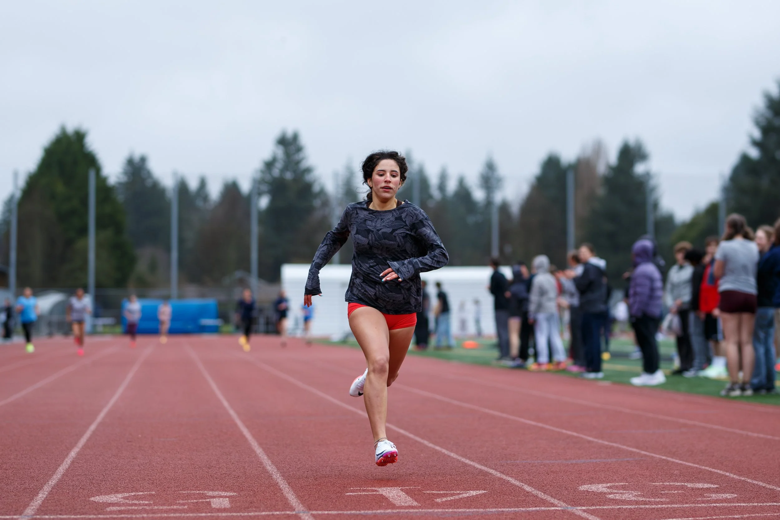 Female athlete wearing dark patterned shirt and red shorts sprints across red track during outdoor race. Spectators watch from sidelines near evergreen trees under overcast sky.