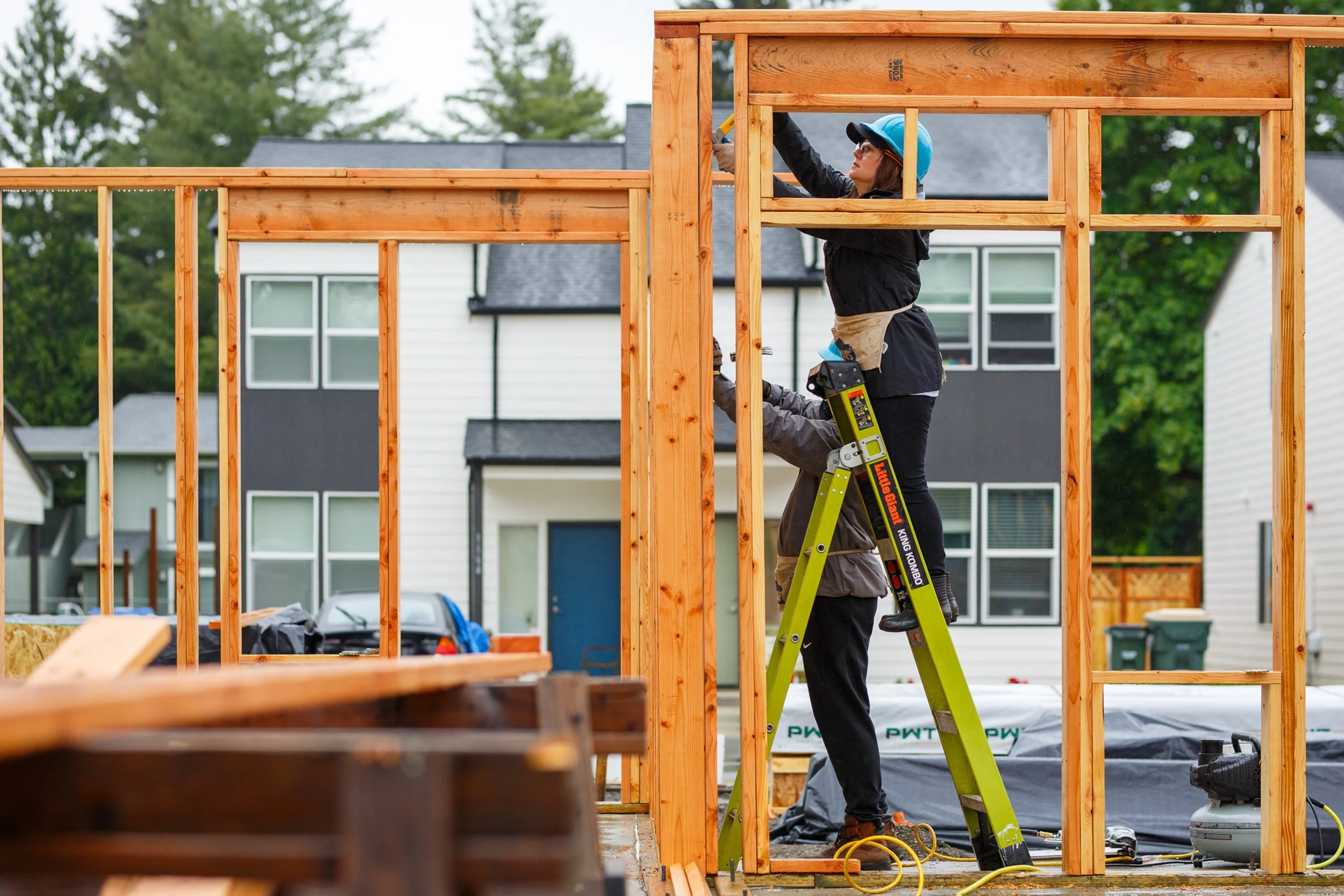 Two volunteers working on a construction site, one on a ladder and one on the foundation, nailing wooden wall framing together with a completed house in the background.