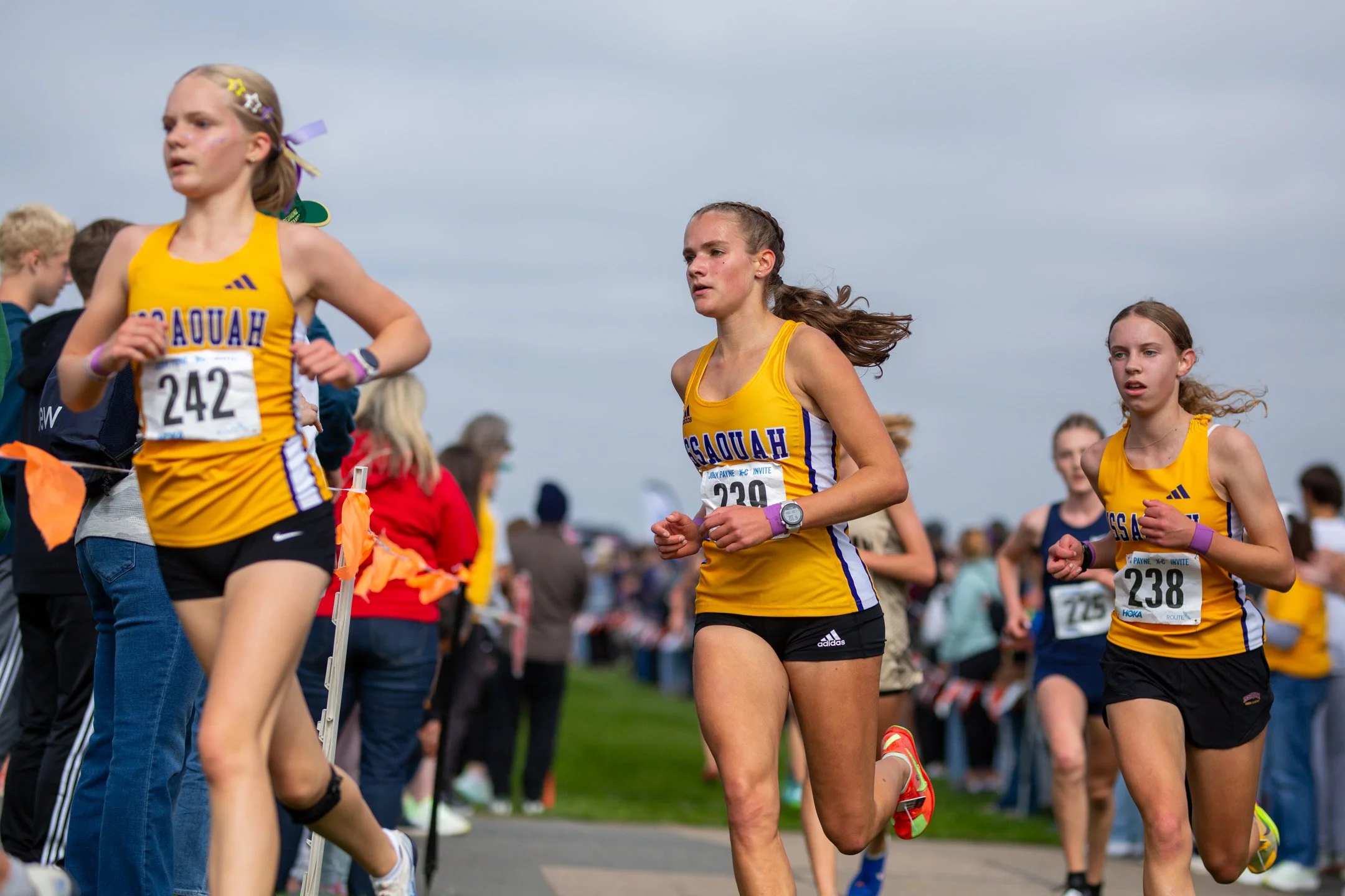 Three Issaquah High School runners in matching gold and black uniforms running side-by-side in tight pack formation past cheering fans.