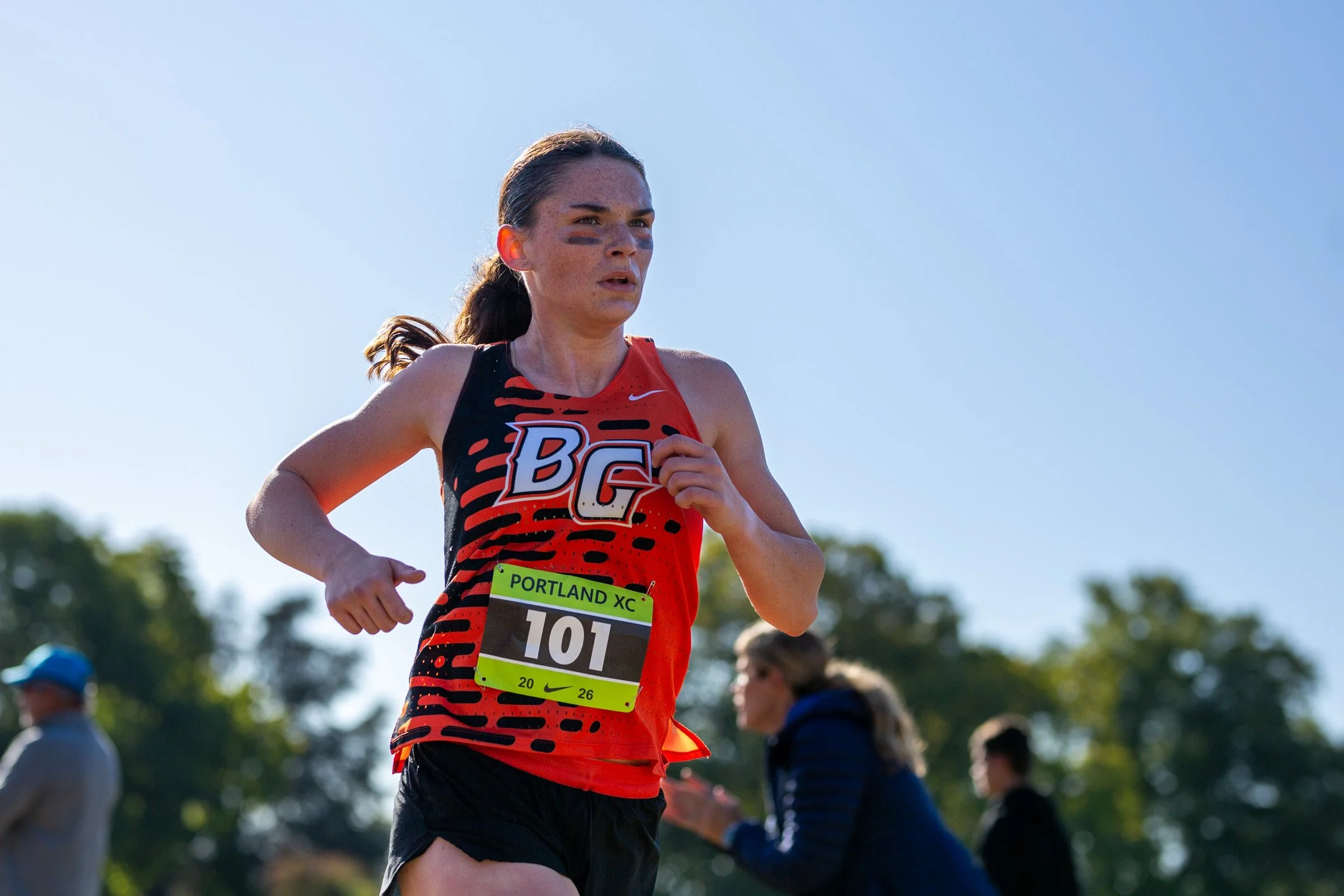 Sophia Rogers of Battle Ground, bib 101, runs in an orange and black “BG” singlet against a blue sky, with spectators and trees behind her.