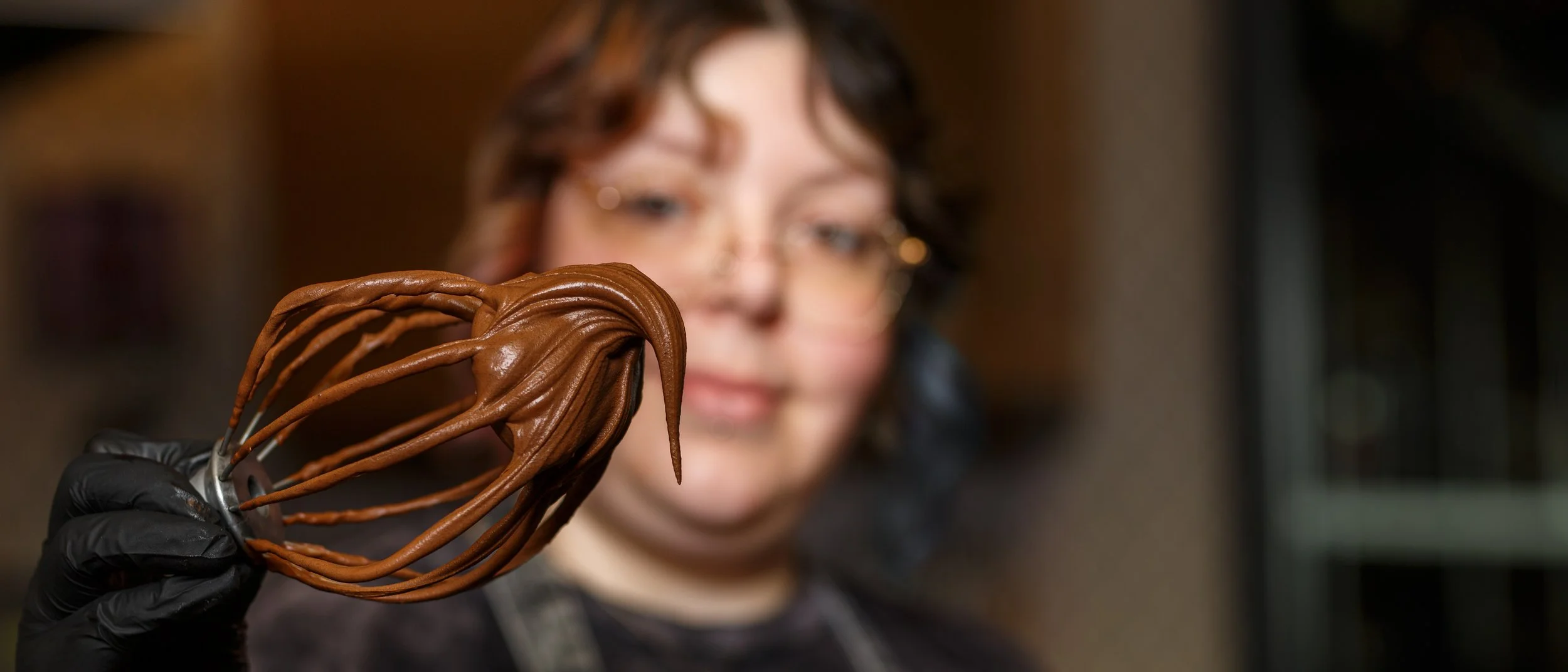 Sharp focus on a large whisk heavily coated in rich chocolate frosting, held by a blurred baker in the background at Tender Velvet Rebel Oven in Olympia, Washington.
