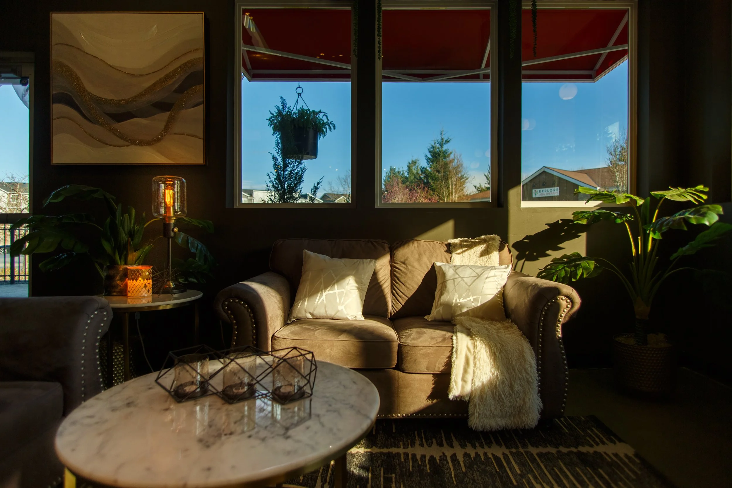 Warm morning sunlight streams through large windows into a cozy cafe seating area featuring a brown velvet loveseat, decorative pillows, potted green plants, and a marble coffee table.