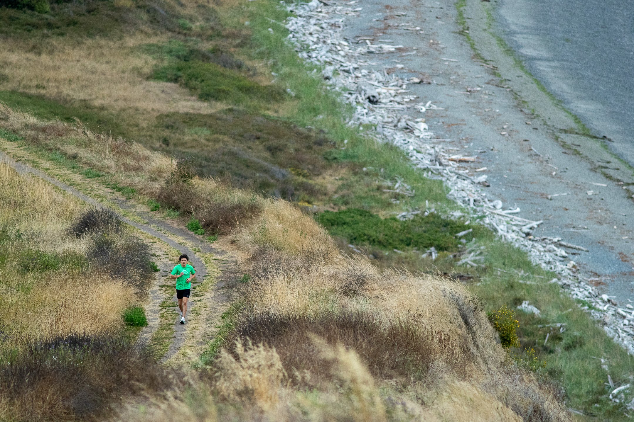 A high-angle view looking down at a high school cross country runner in a bright green shirt running along a dirt trail on the coastal bluffs above a driftwood-lined shore.