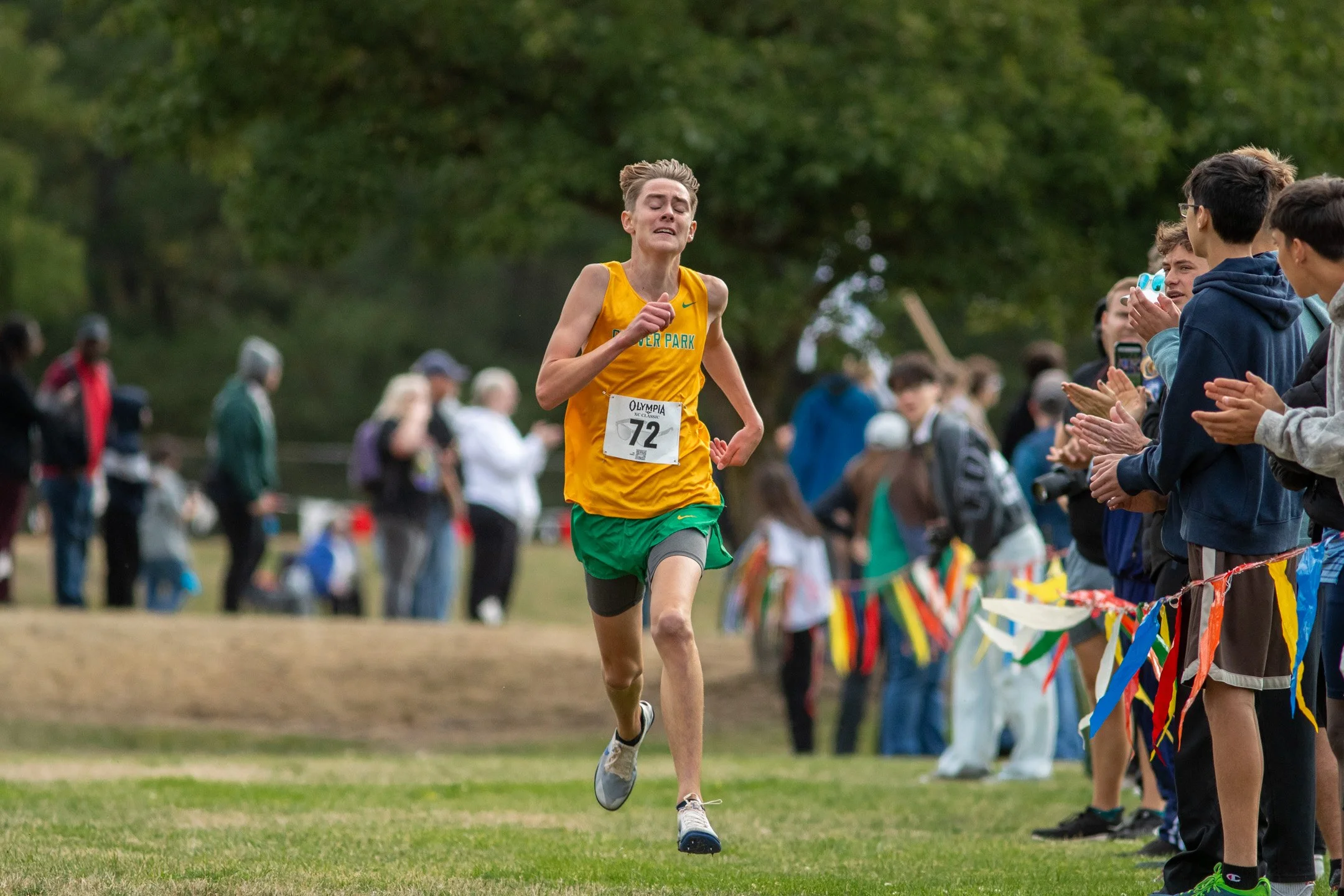 Boy in yellow and green Clover Park jersey, bib 72, running alone past a flag-lined finishing chute.