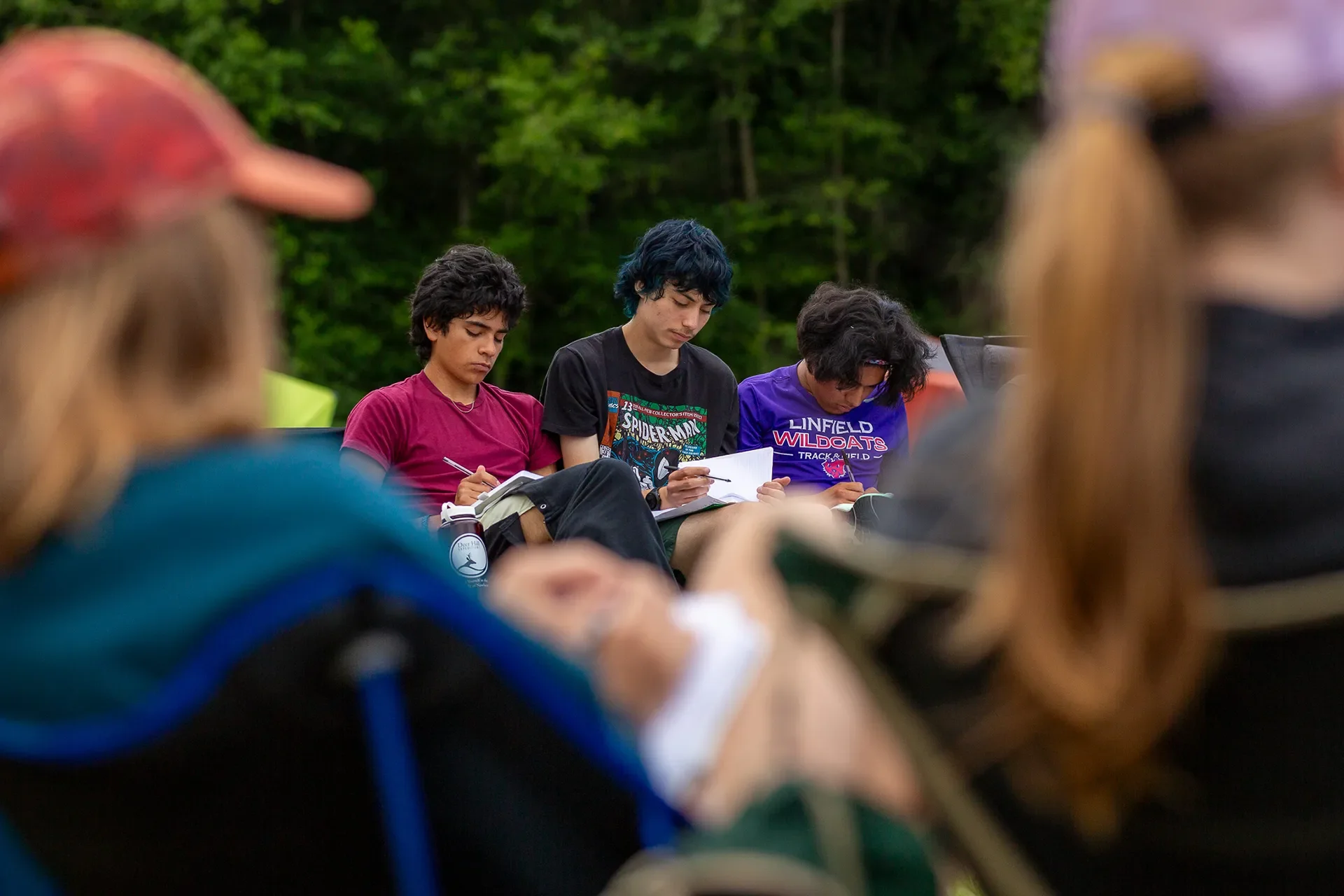 Three high school boys sitting in camping chairs outdoors, writing in notebooks with a blurred foreground.