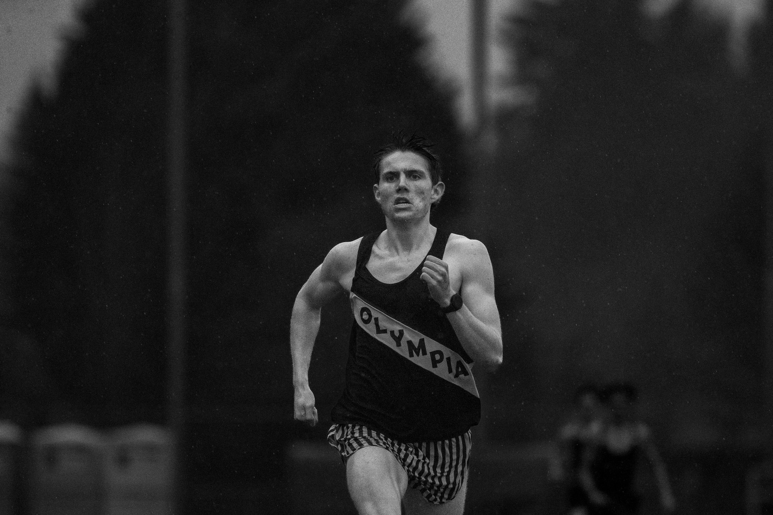 Gritty black and white action shot of Olympia High School track athlete Caden Durocher sprinting alone toward the camera through a heavy rainstorm during the final stretch of the 1600m race.