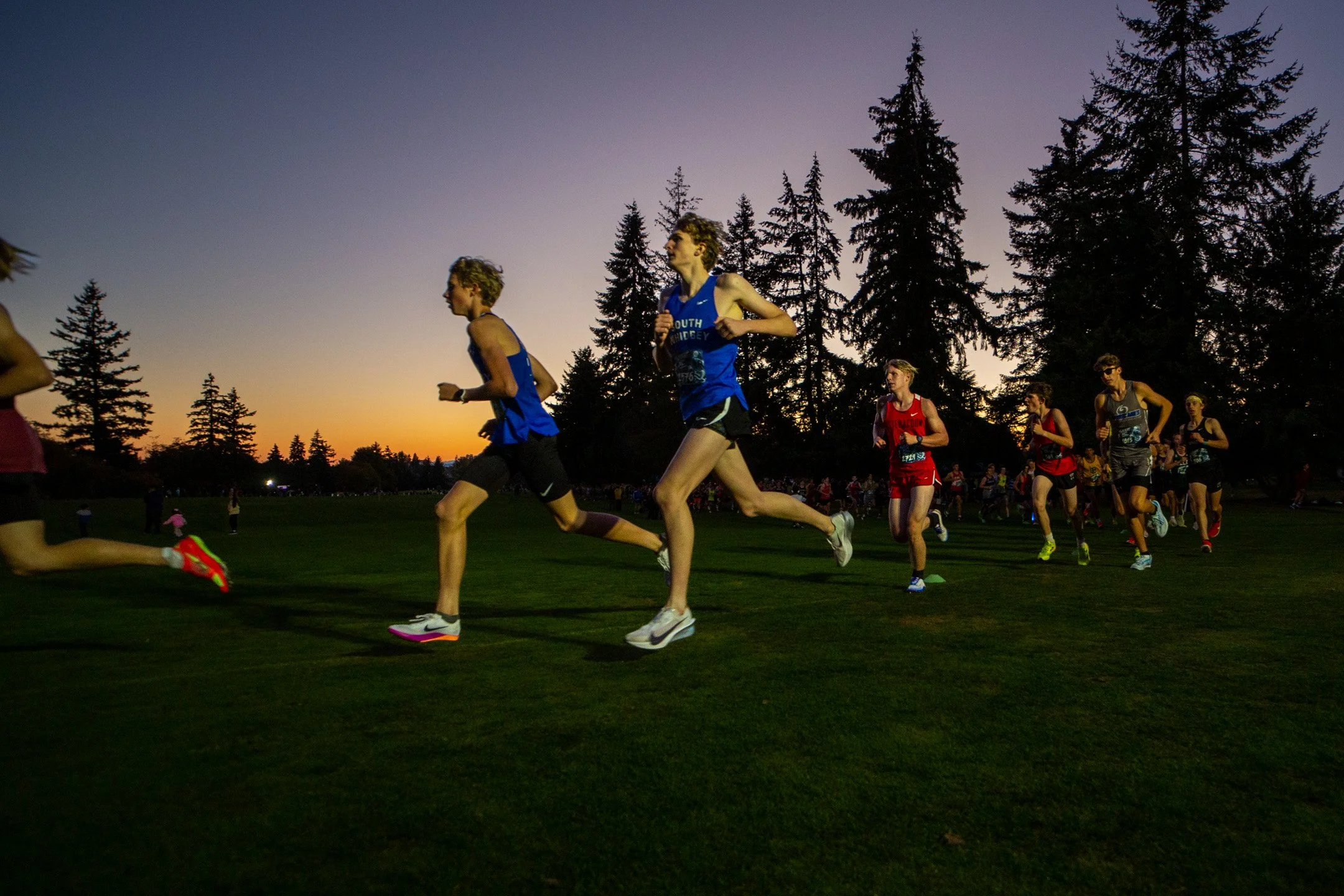 High school boys cross country runners racing across a golf course fairway under a vivid orange and dusky purple sunset sky bordered by tall evergreen trees.