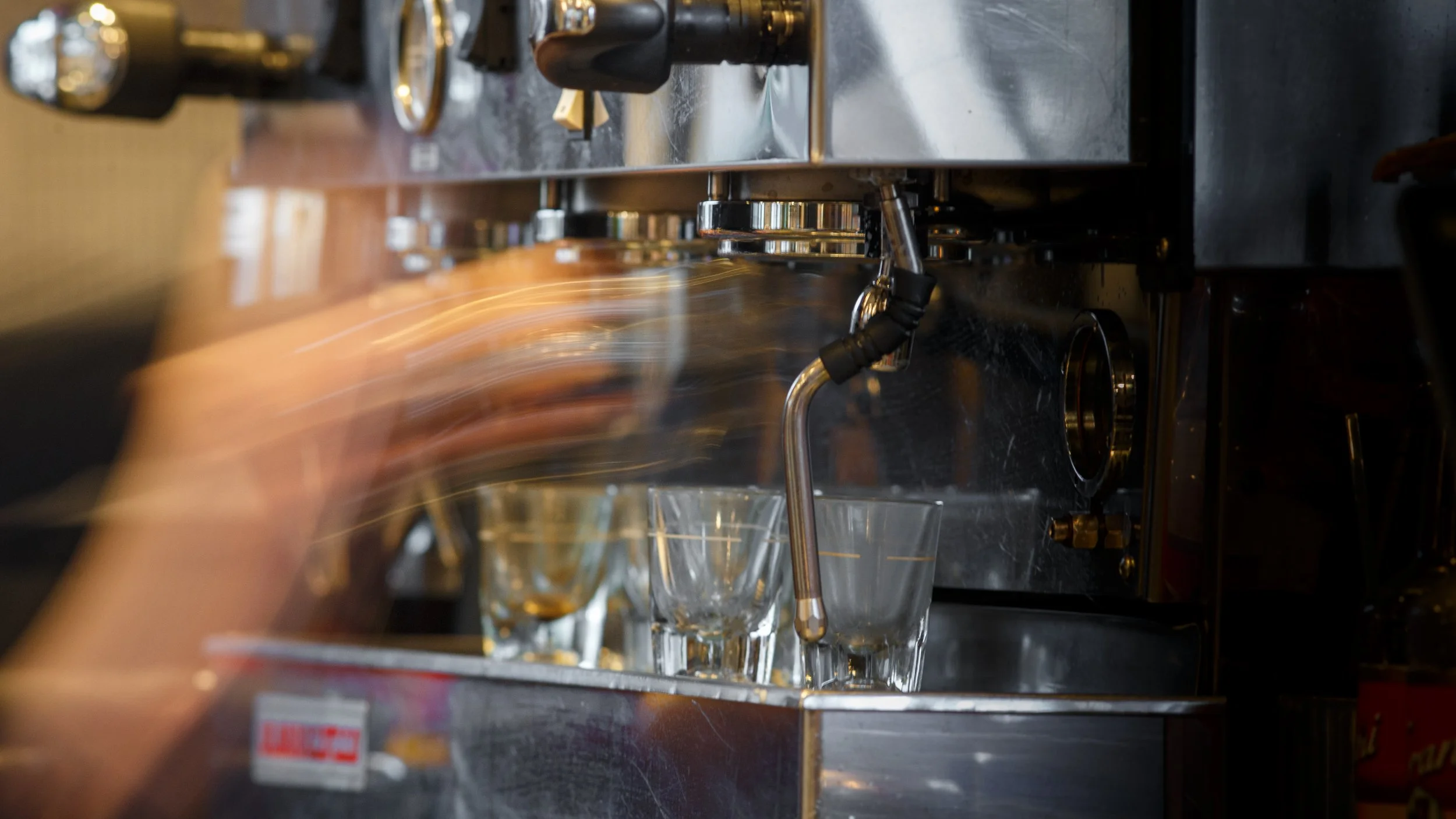 A barista's arm creates a fast motion blur while operating a shining stainless steel espresso machine with small glass cups at Taylor Rays Cafe in Olympia Washington.