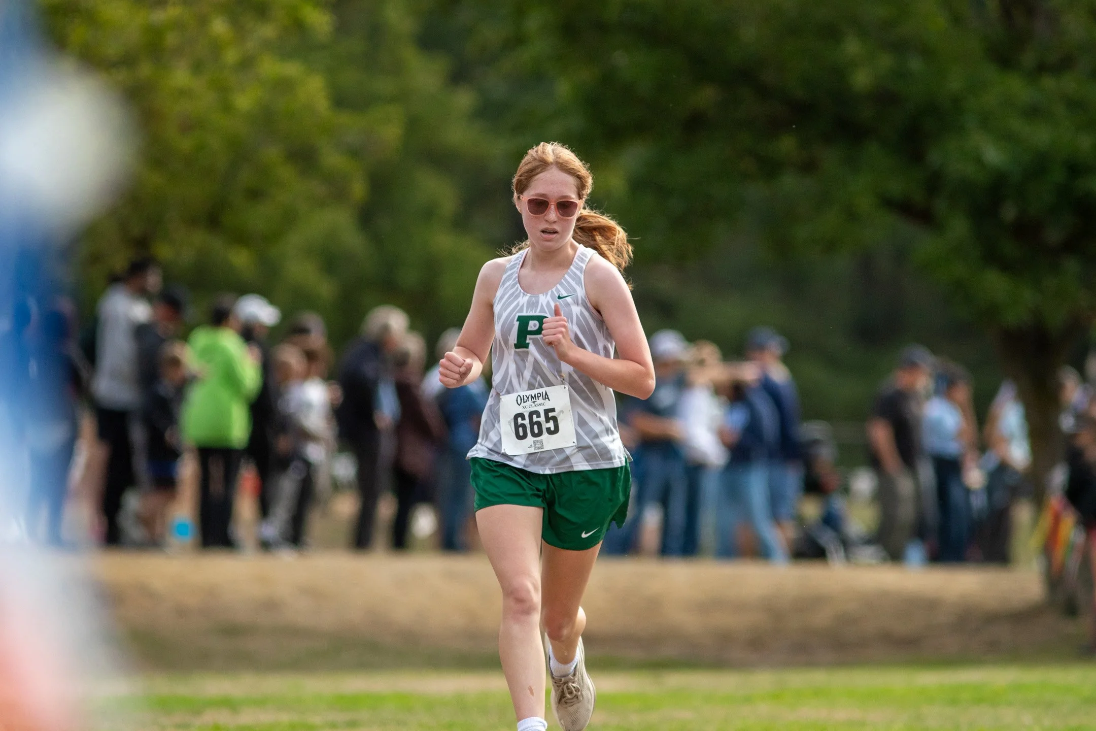 Girl in white and green Peninsula jersey, bib 665, running past spectators lining the course.