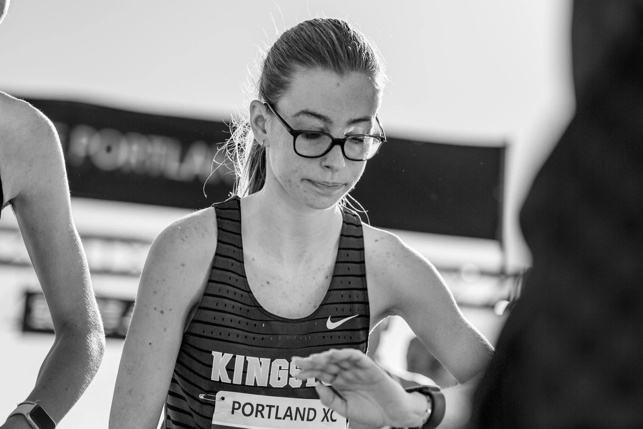 Black-and-white image of Kingston senior Emma Grice in glasses, looking down after the finish with a hand at her wrist.