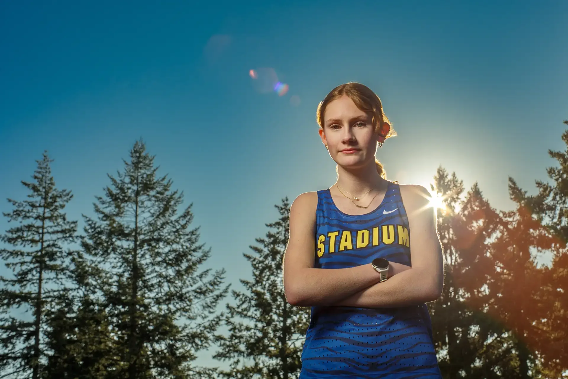 Portrait of a female runner with arms crossed standing on a track with the sun creating a starburst flare behind her shoulder through the trees.
