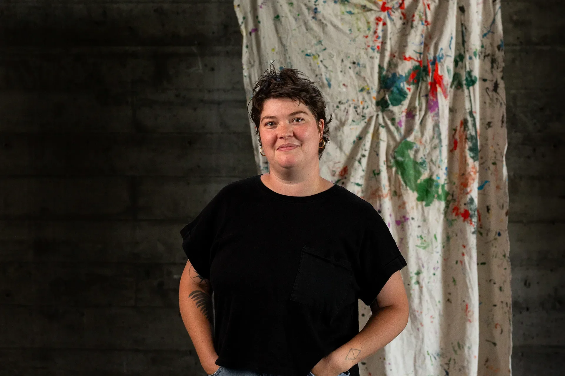 Artist Fern Tallos smiles in a black shirt before a paint-streaked drop cloth and brick wall.