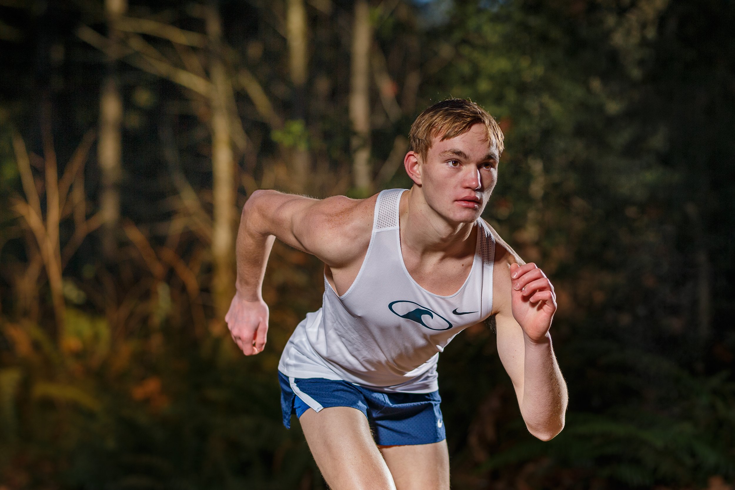 Action shot of a male cross country runner sprinting through a dark forest trail, illuminated by dramatic off-camera flash.