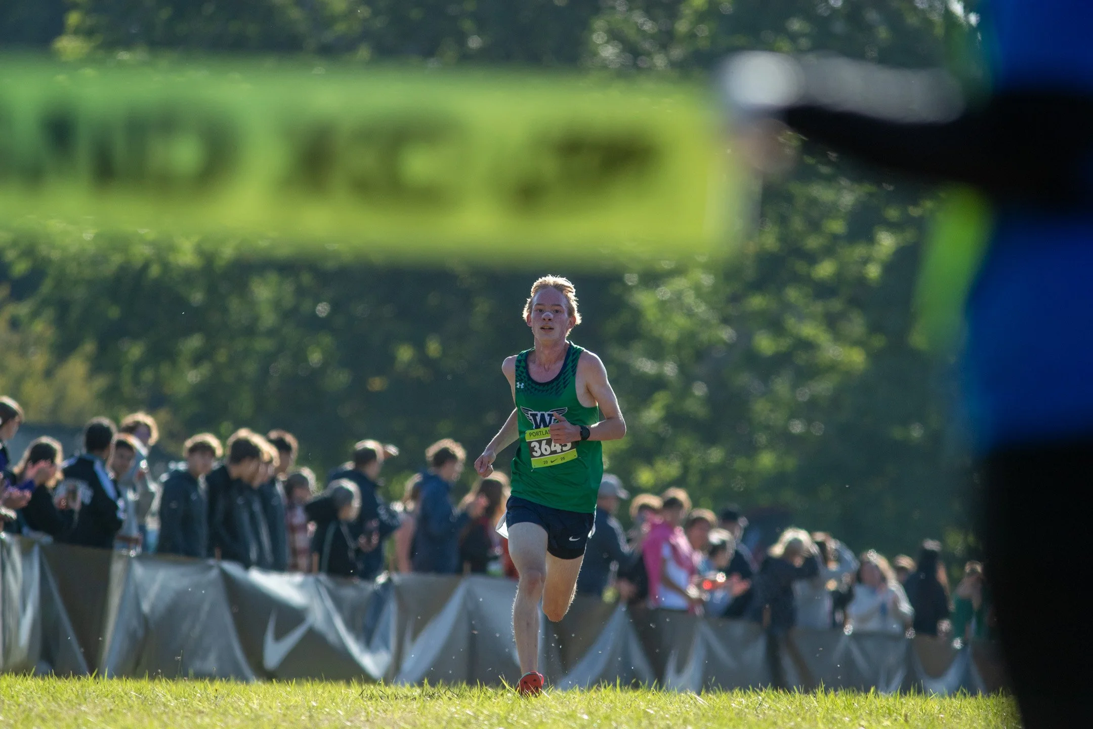 Woodinville sophomore Oskari Vuorela in a green singlet runs toward the camera, a blurred yellow finish tape in the corner of the frame.