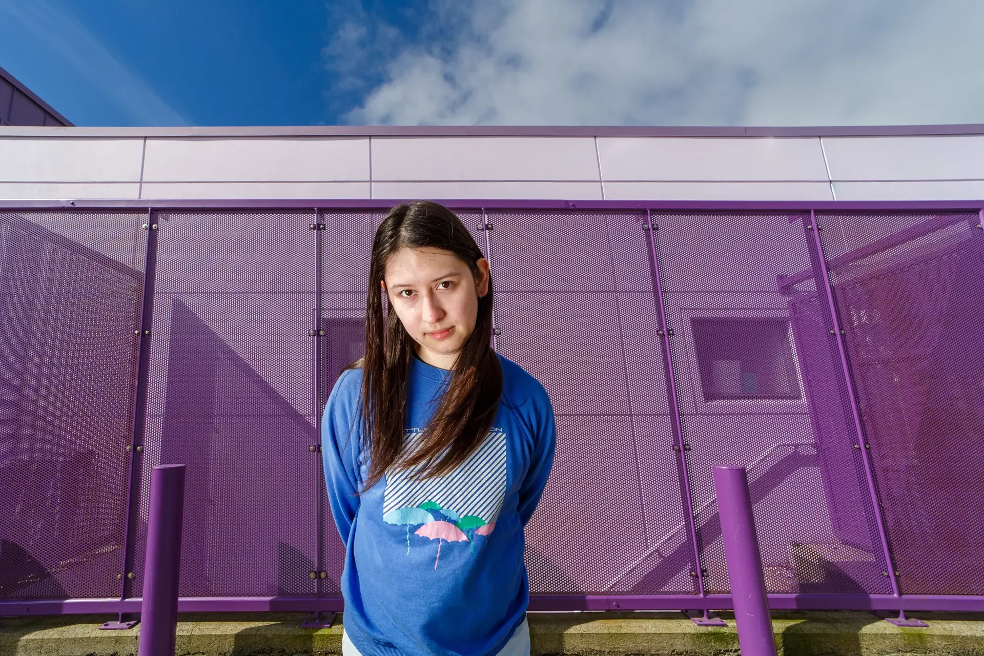 Local Olympia comedian Nat Penrose wearing a blue graphic sweater, giving a serious, deadpan expression while standing in front of a vibrant purple perforated metal building.