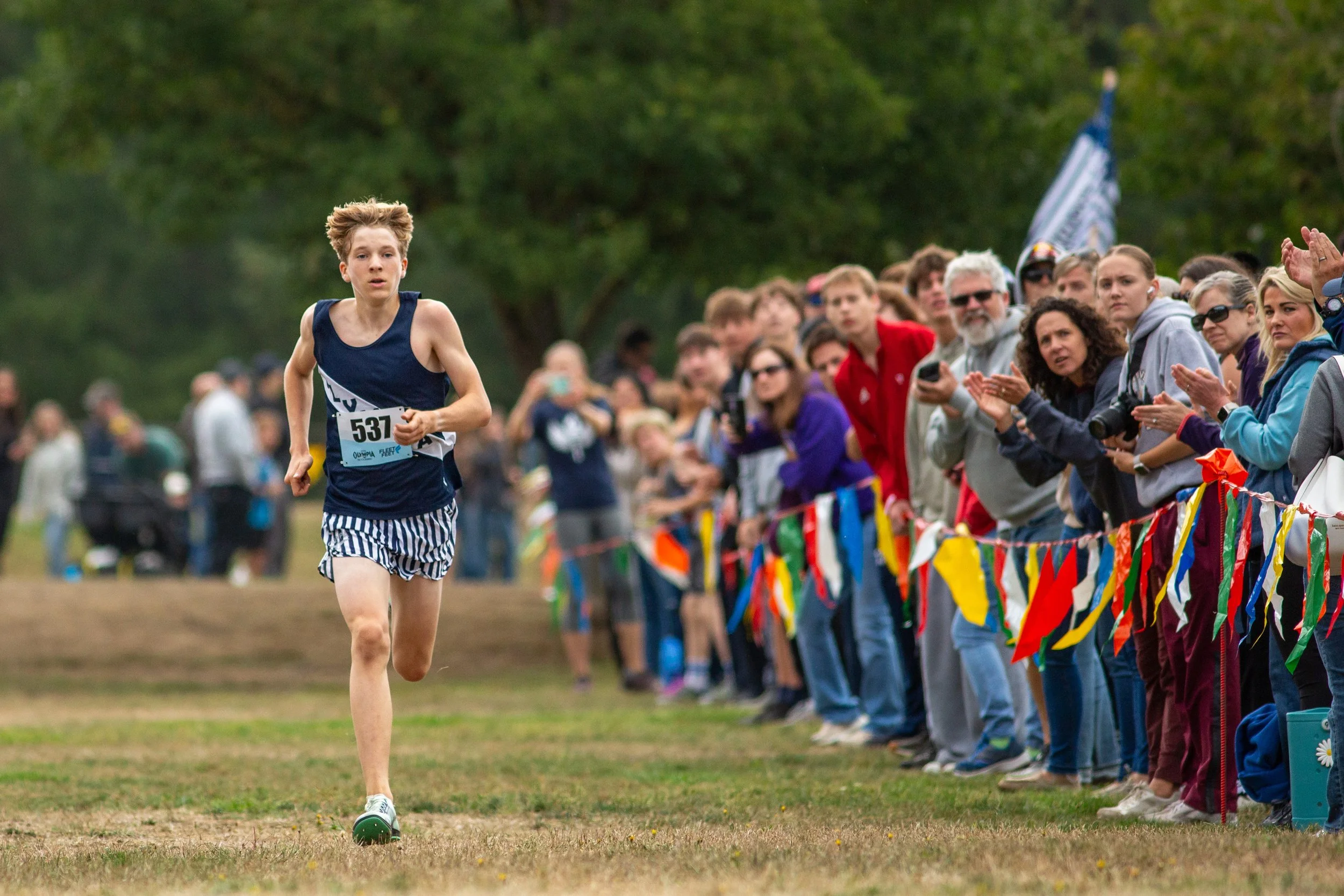 Boy in navy Olympia singlet and zebra-print shorts running alone along a flag-lined finishing chute.