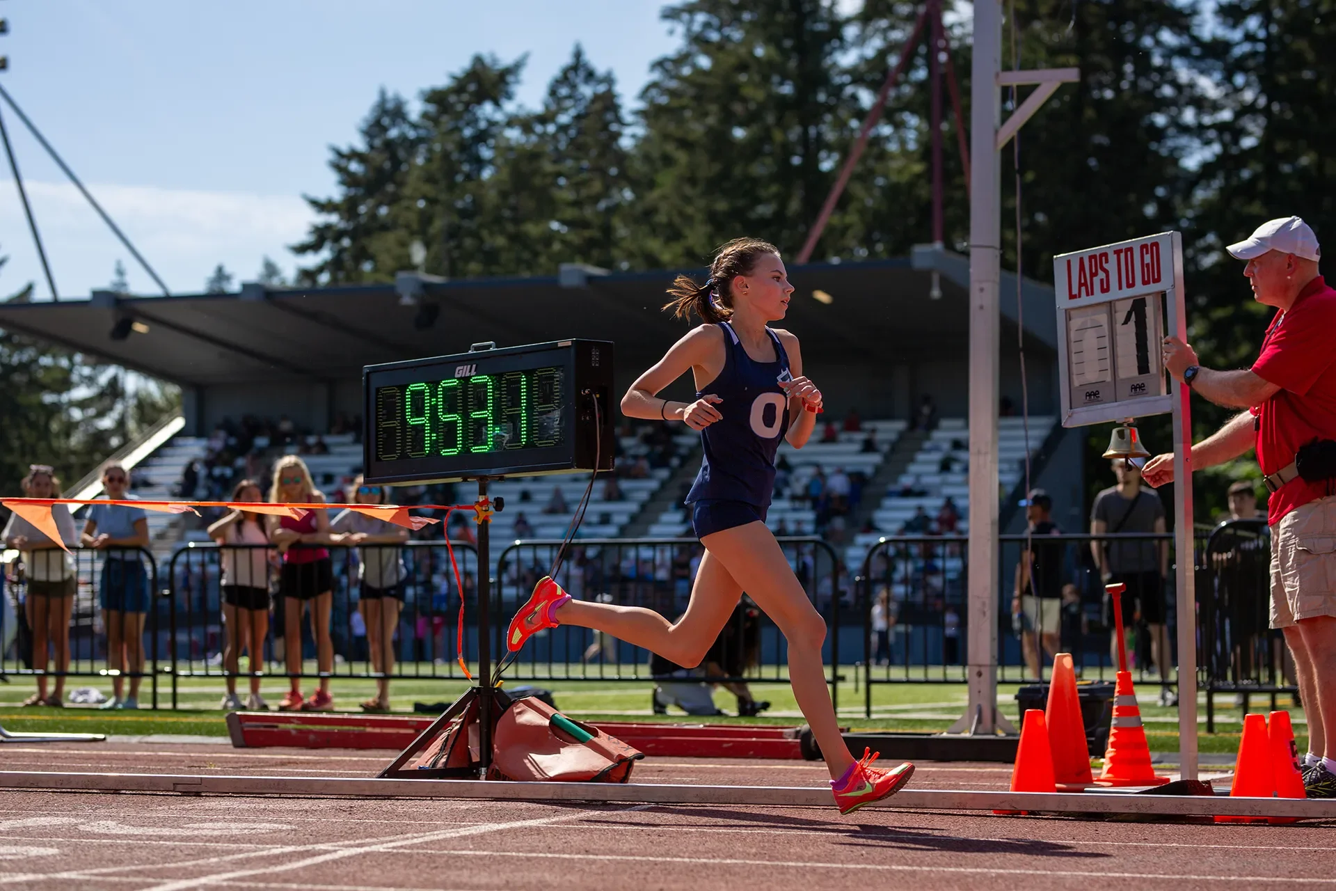 Olympia High School runner Leah Hennen in a navy blue singlet and bright pink shoes passing an official holding a lap counter showing "1" lap to go during the 3200m race.