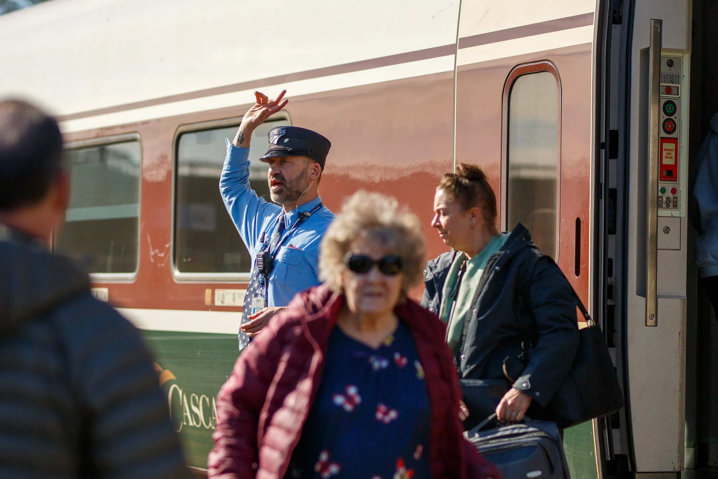 A uniformed Amtrak conductor signals with his hand as passengers board an Amtrak Cascades train at the Olympia / Lacey Amtrak station, photographed by Russell Moore.