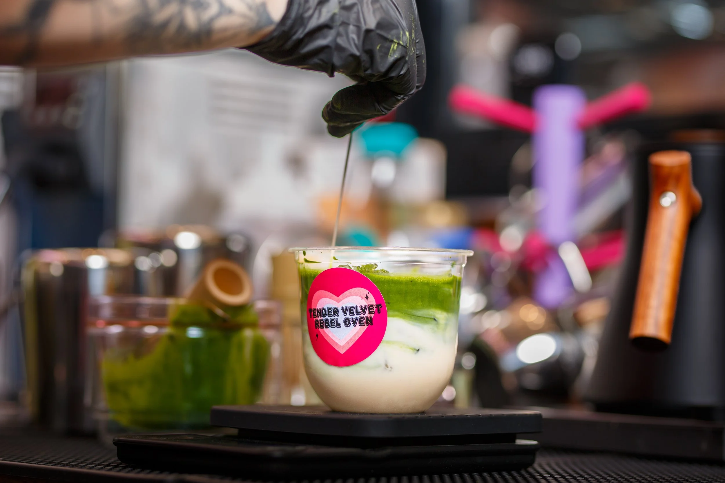 A close-up shot of a barista in black gloves mixing a vibrant green iced matcha latte in a plastic cup featuring a pink Tender Velvet Rebel Oven heart logo sticker.