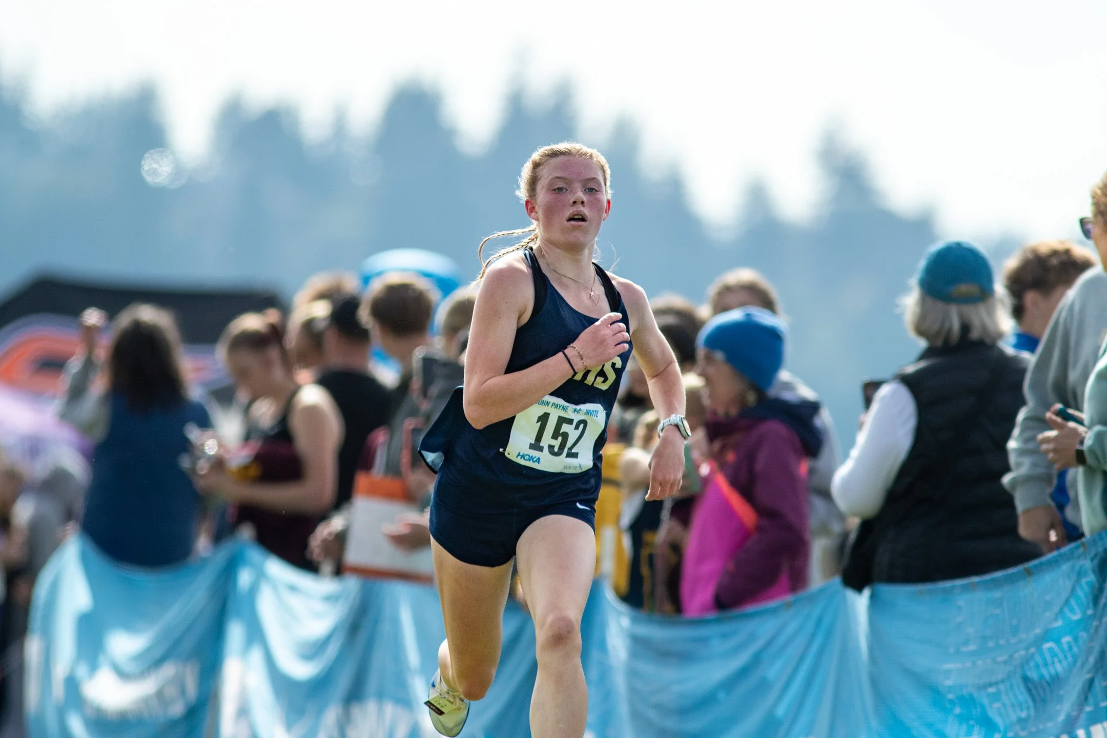 Arlington freshman Ellie Oakes in a navy blue jersey with bib 152 driving toward the finish line past cheering spectators.