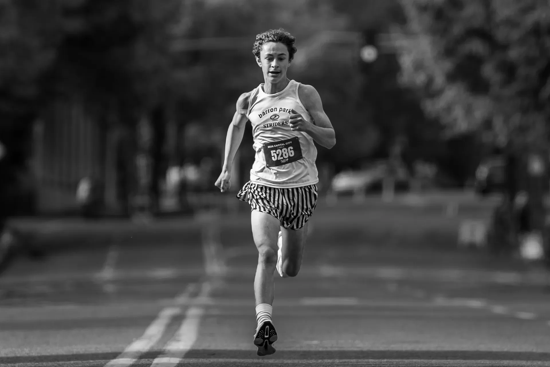 Black and white action photo of youth runner Quin Zola sprinting alone down the road during the 2026 Capital City 5-Mile race.