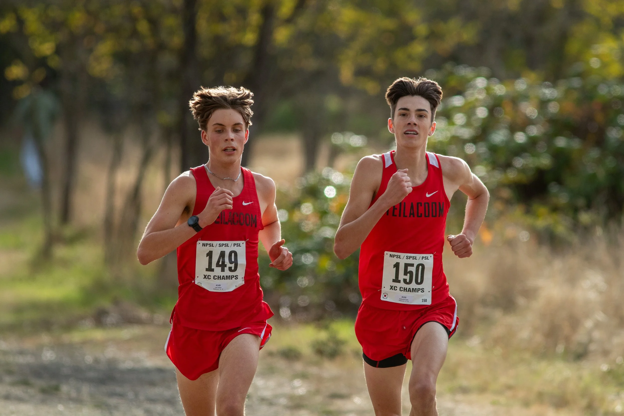 Zechariah Swift and Luke Slater of Steilacoom, bibs 149 and 150, run side by side in red singlets on a dirt course lined with brush.
