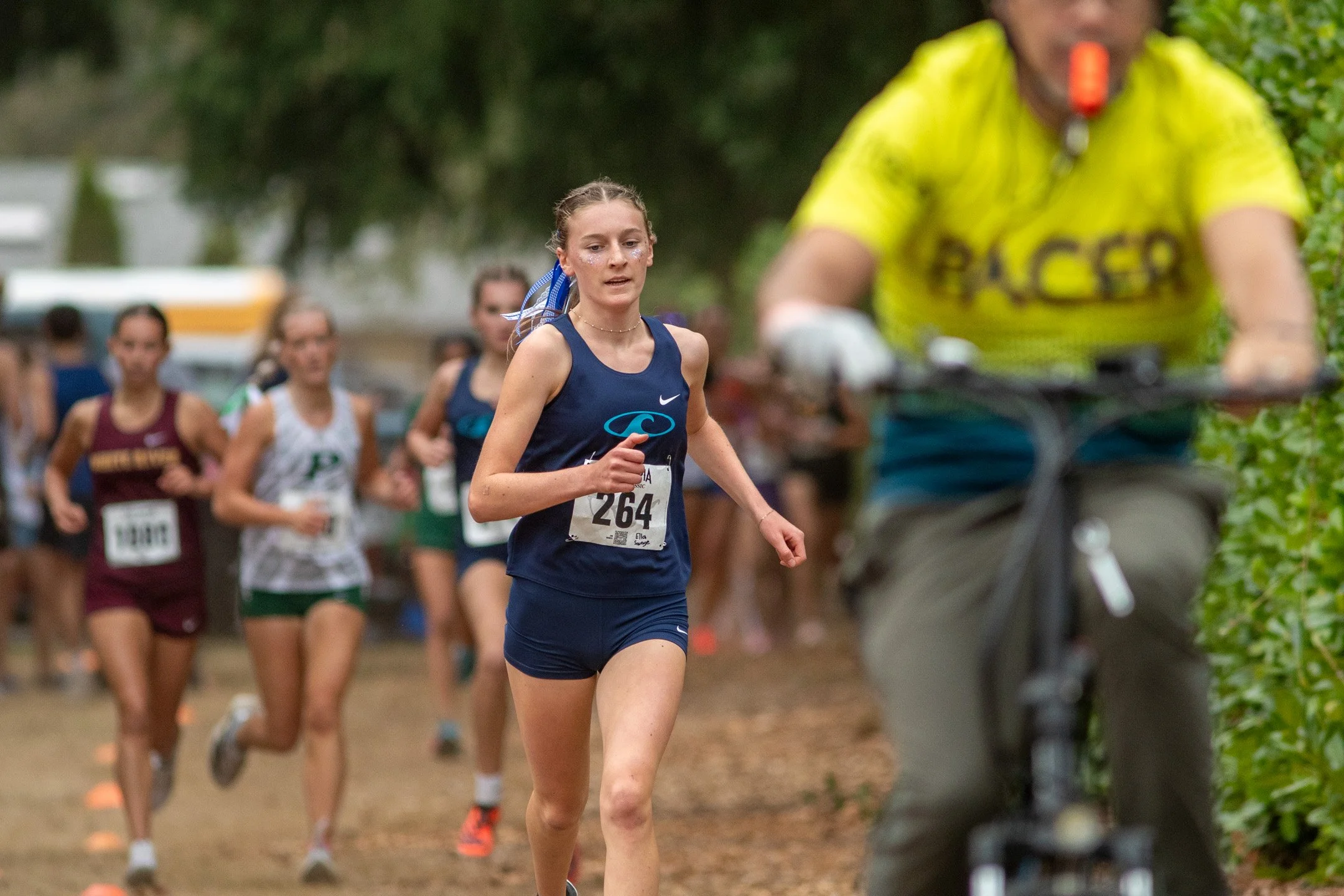Girl in navy Gig Harbor jersey, bib 264, leading a pack of junior girls runners past an official pace escort on a bicycle.